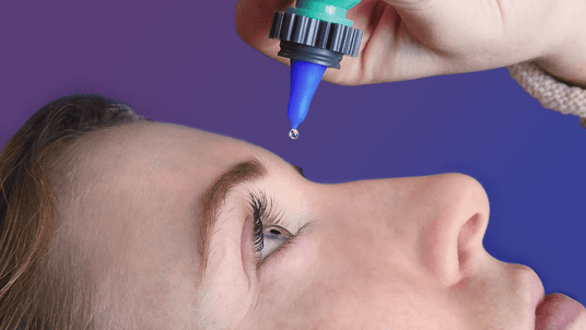 Close-up of a person lying down, looking up as a dropper releases an eye drop into their right eye. The background is a gradient of purple and blue.