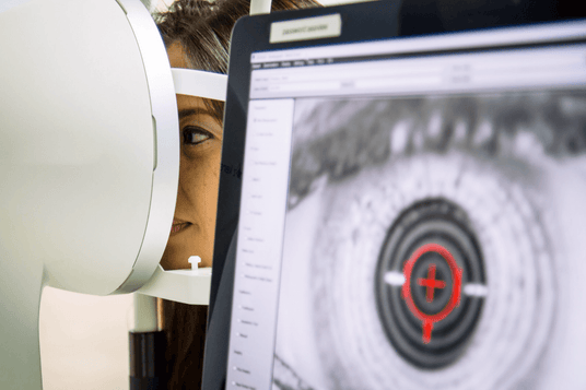 A person looking into an eye examination device, with a focus on their eye. In the foreground, a computer screen shows a close-up image of an eye with a target-like graphic overlay.