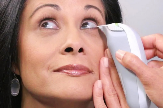 A person holds a handheld device near a womans eye, pressing gently on her lower eyelid. The woman appears calm, with makeup and earrings visible, as she looks upwards. The background is a soft grey.