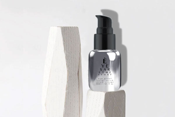 A sleek, metallic bottle with a black pump dispenser is displayed on geometric white wooden blocks against a light gray background. The bottle is labeled Eye Serum.