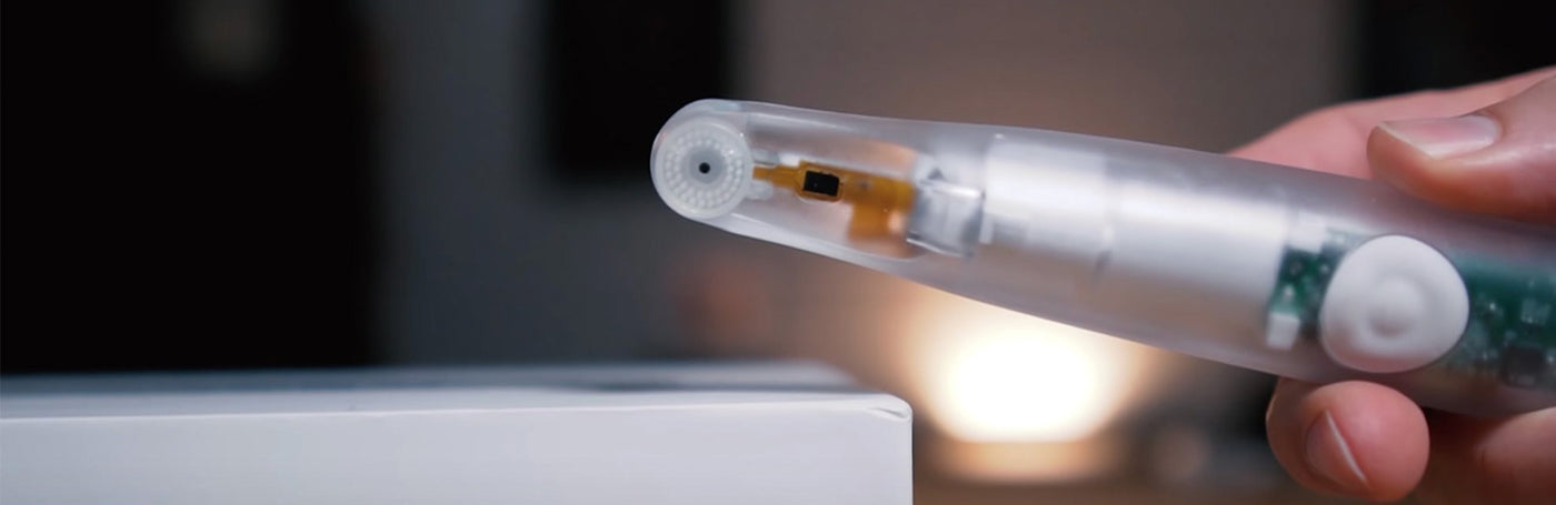Close-up of a person holding a translucent electronic pen device, with visible internal components. The pen is angled towards a white box on a blurred background.