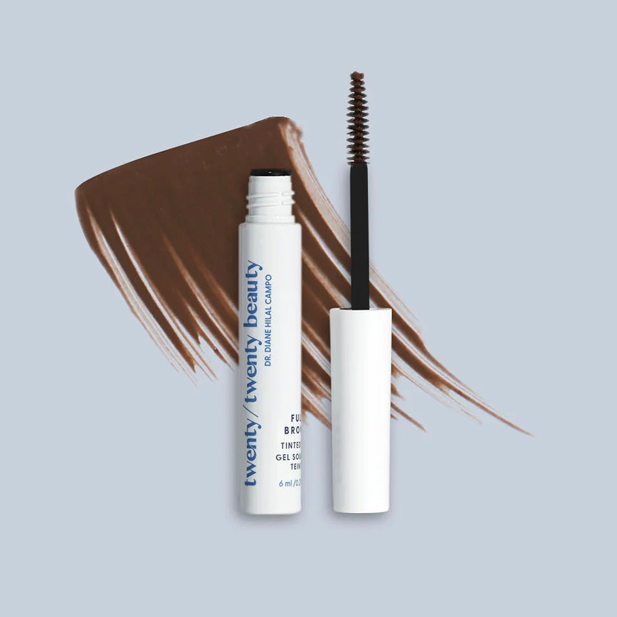A tube of Twenty Twenty Full Brows Tinted Gel rests on a gray surface with its open wand. Brown gel is smeared behind it, showcasing its color for fuller eyebrows. The vegan and cruelty-free formula is in a white tube with blue text, containing 6ml of product.