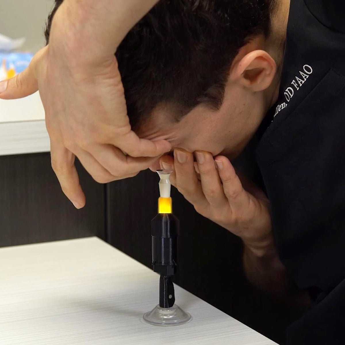 Using the Augmented Vision Labs S5 Mini Inserter for Scleral & Hybrid Lens Insertion, a person leans over a table with their head tilted. The hands-free lens stabilization device, upright on a suction base, ensures precise alignment and ease. Free video training is included.