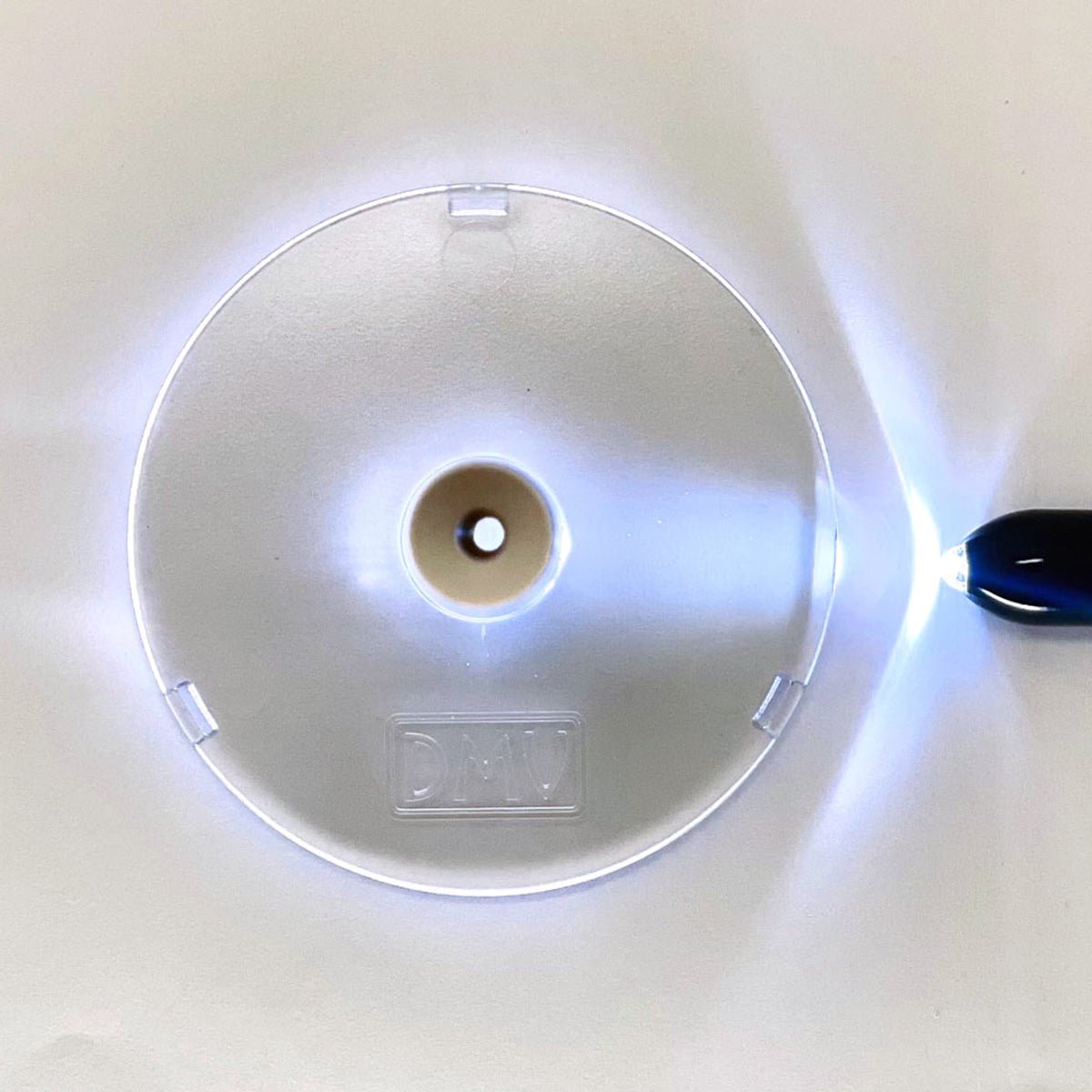 A circular, translucent plastic scleral lens inserter by DMV has a center hole and is set on a white background. A light from the right casts a glow around it. Embossed with DMV, it emphasizes its Made in USA quality.