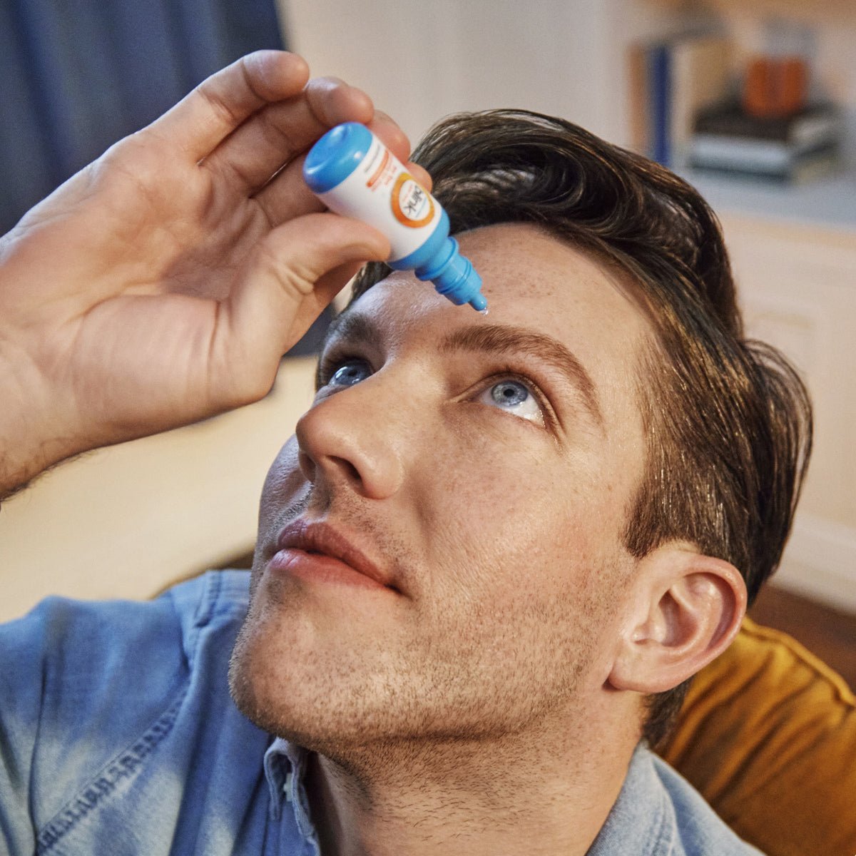 In a well-lit indoor area with blurred furniture and curtains, a person in a blue shirt carefully tilts their head back to apply Blink Triple Care 3 in 1 Eye drops by Bausch + Lomb for dry eyes, holding the small bottle with precision.
