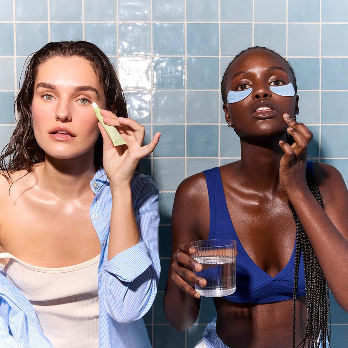 Two women in a bathroom apply skincare against a blue tiled wall. The woman on the left uses Daily Practice Firm & Restore Eye Cream – Smooth, Hydrate & Lift (15mL), while the woman on the right holds a glass of water with eye patches to reduce fine lines.