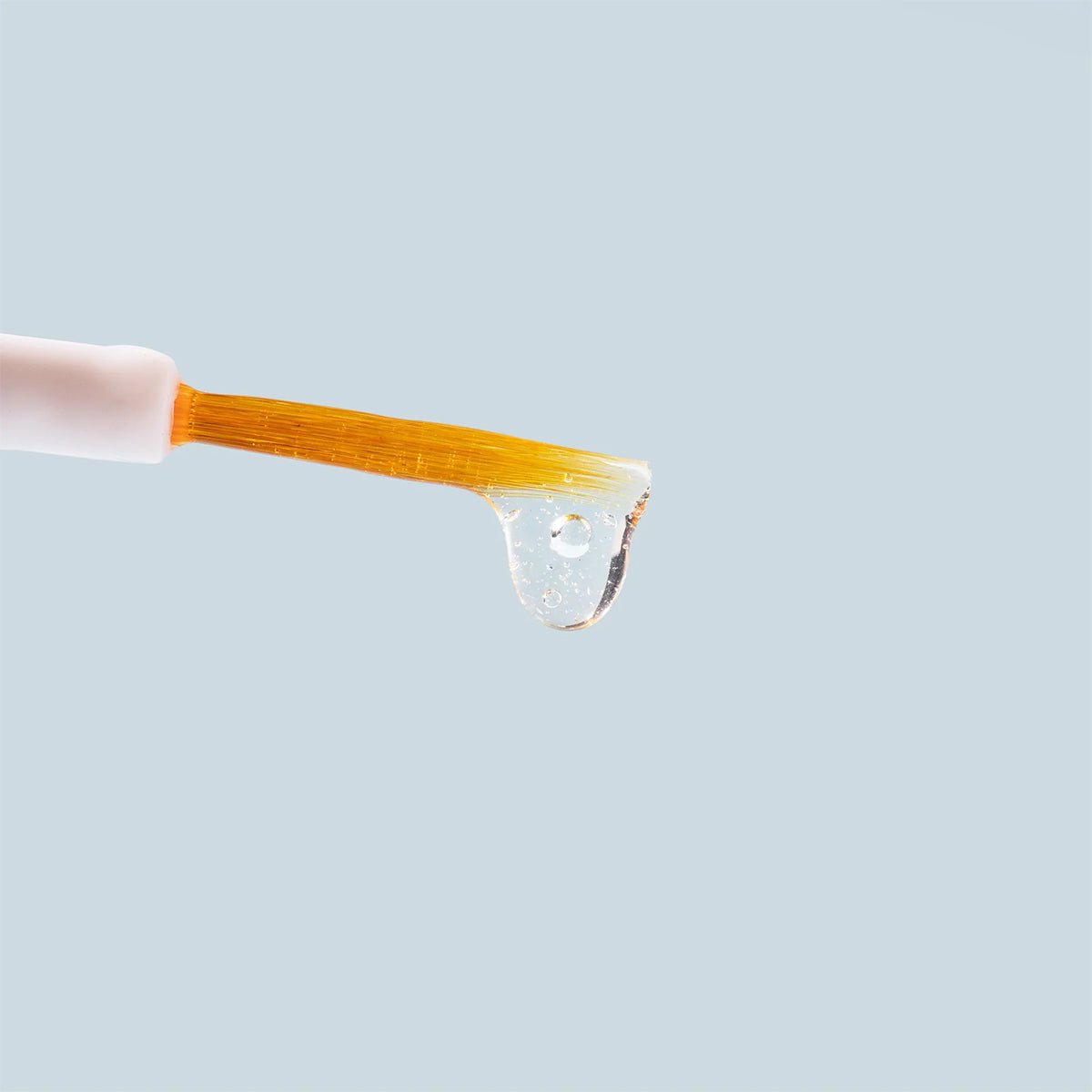A close-up of an orange-bristled brush with clear liquid on its tip, set against a light blue background, depicts the Daily Practice Lash Enhancing Serum (4ml) with a white handle that promises fuller, longer, healthier eyelashes.