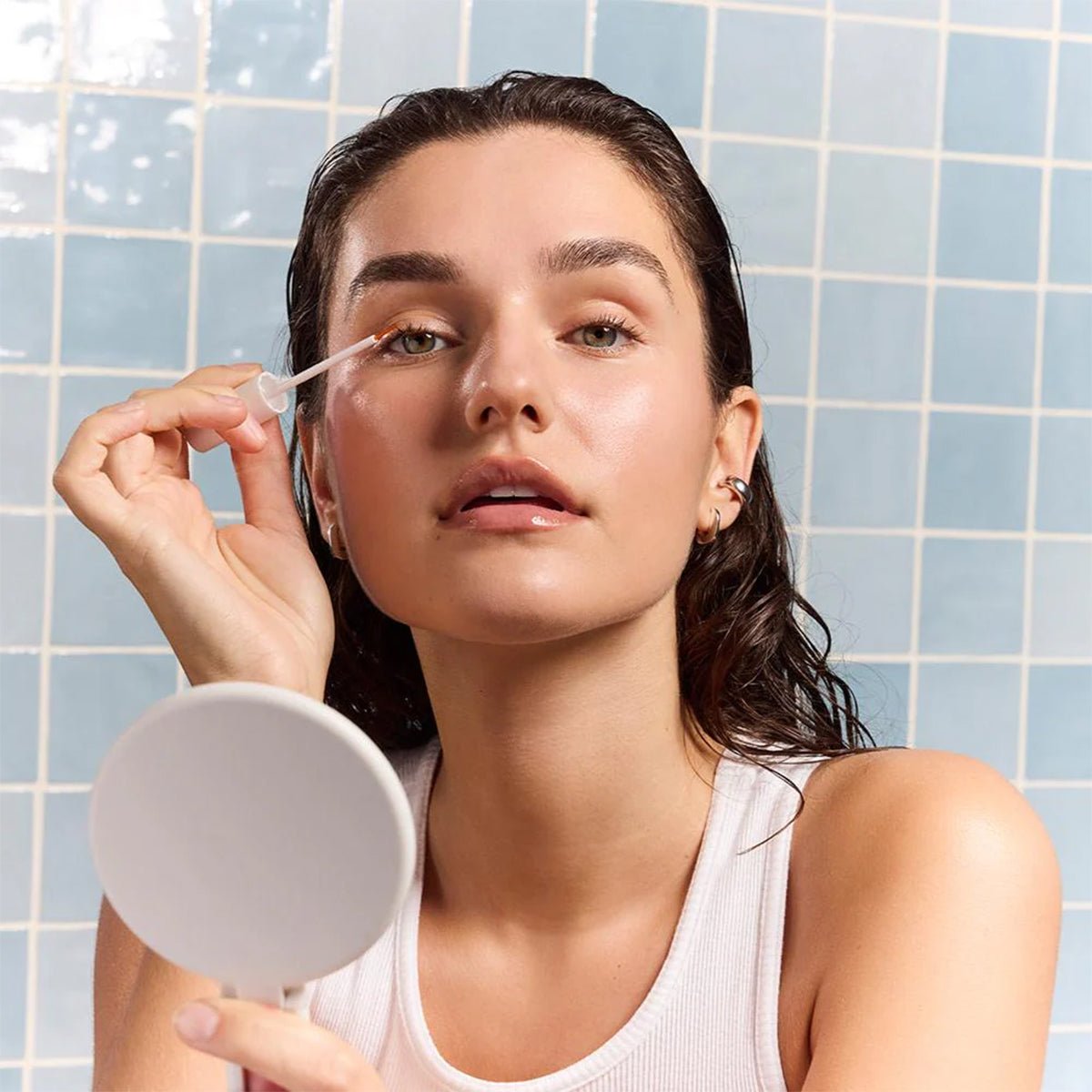 A person with wet hair is applying Daily Practice Lash Enhancing Serum using a dropper while holding a small mirror. They are wearing a white tank top and standing in front of a tiled bathroom wall.
