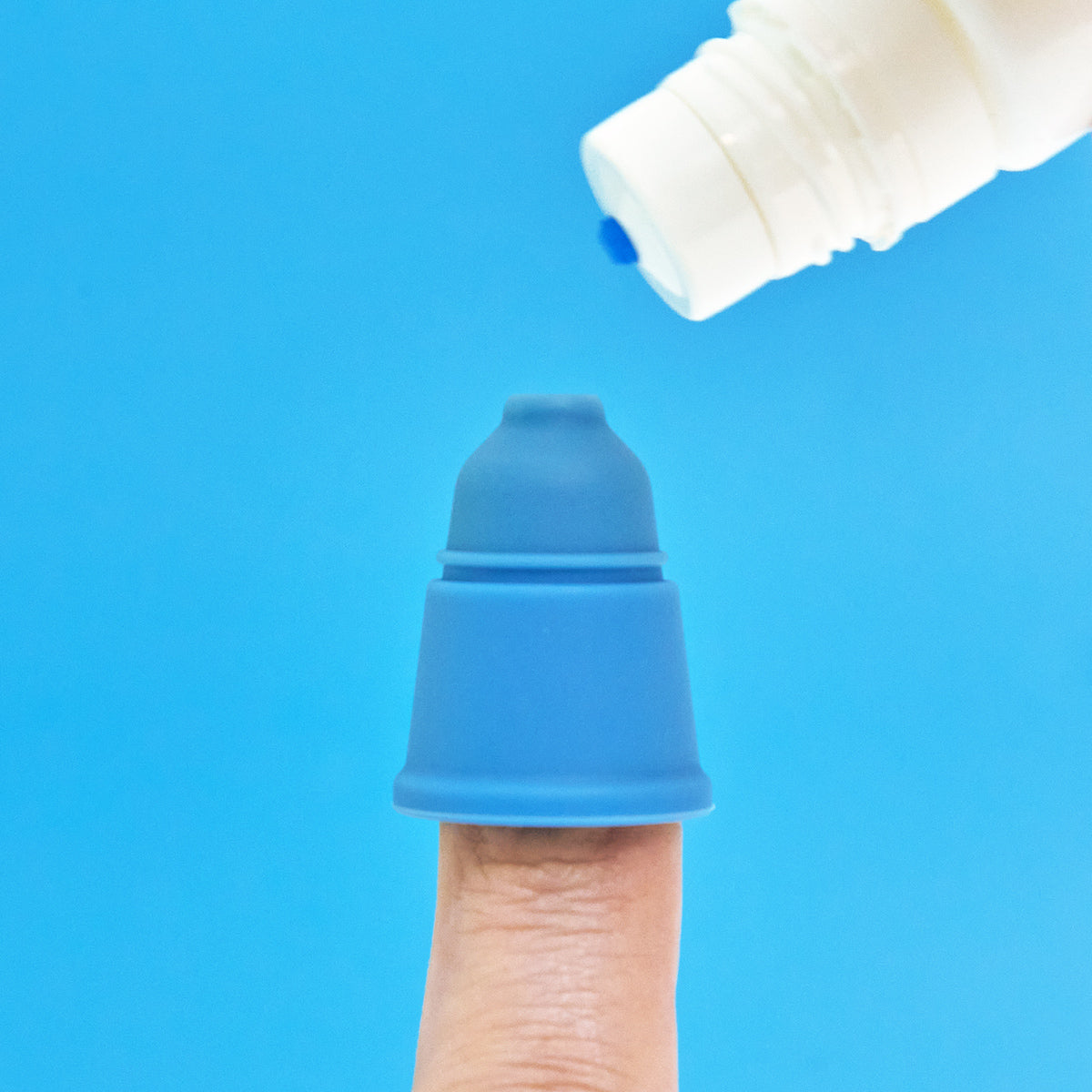 A finger shows a small blue Magic Touch Eye Drop Applicator silicone cup, topped with a white bottle with a blue cap against a solid blue background.