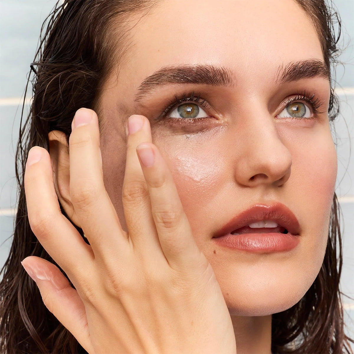 With wet hair and minimal makeup, the person applies Daily Practice Nourishing Makeup Melting Balm to their cheek in an upward motion. They gaze upward, displaying clear skin and well-defined eyebrows in a bright setting.