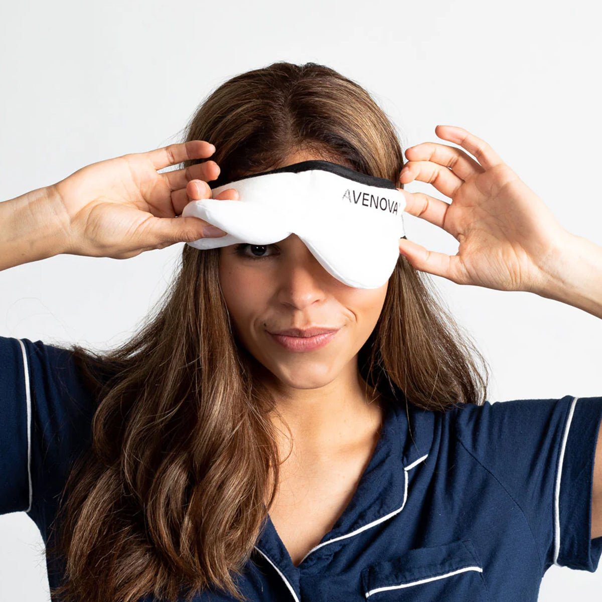 A woman with long brown hair wears a navy blue pajama top and smiles as she adjusts the Avenova Microwavable Heat Eye Compress Mask by NovaBay over her eyes, providing relief from blepharitis and dry eye, against a plain white background.