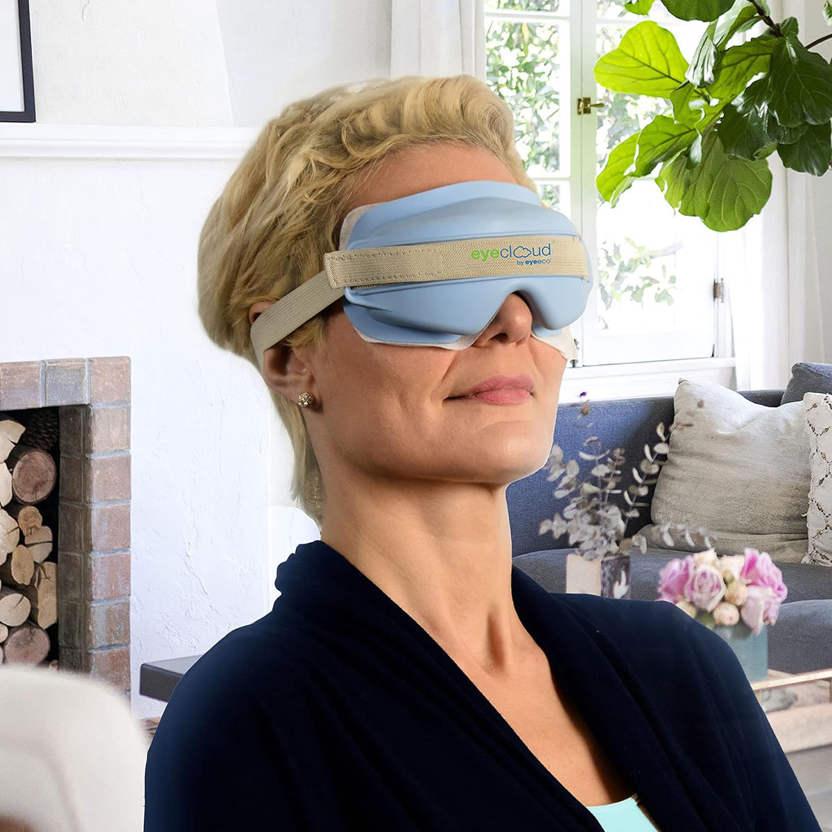 A person with short blonde hair relaxes in a living room, wearing a blue EyeCloud Home Treatment Kit by PRN, known for soothing dry eyes. The space features a cozy fireplace with logs, a large plant, and flowers on the table.