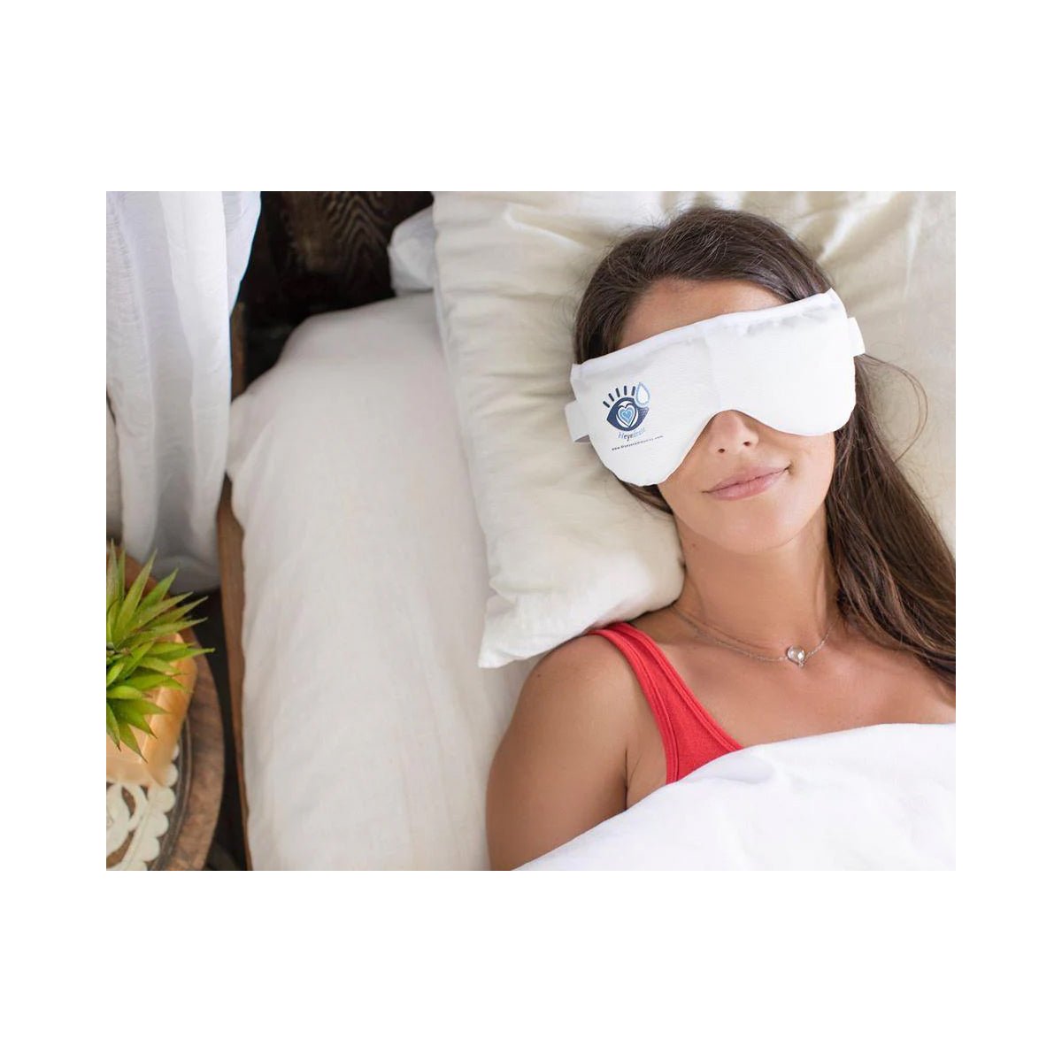A person in a red top lies in bed with a PRN Heyedrate Warm Compress Eye Mask, enjoying dry eye relief while covered by a white blanket. Nearby, a wooden side table holds a small potted plant.