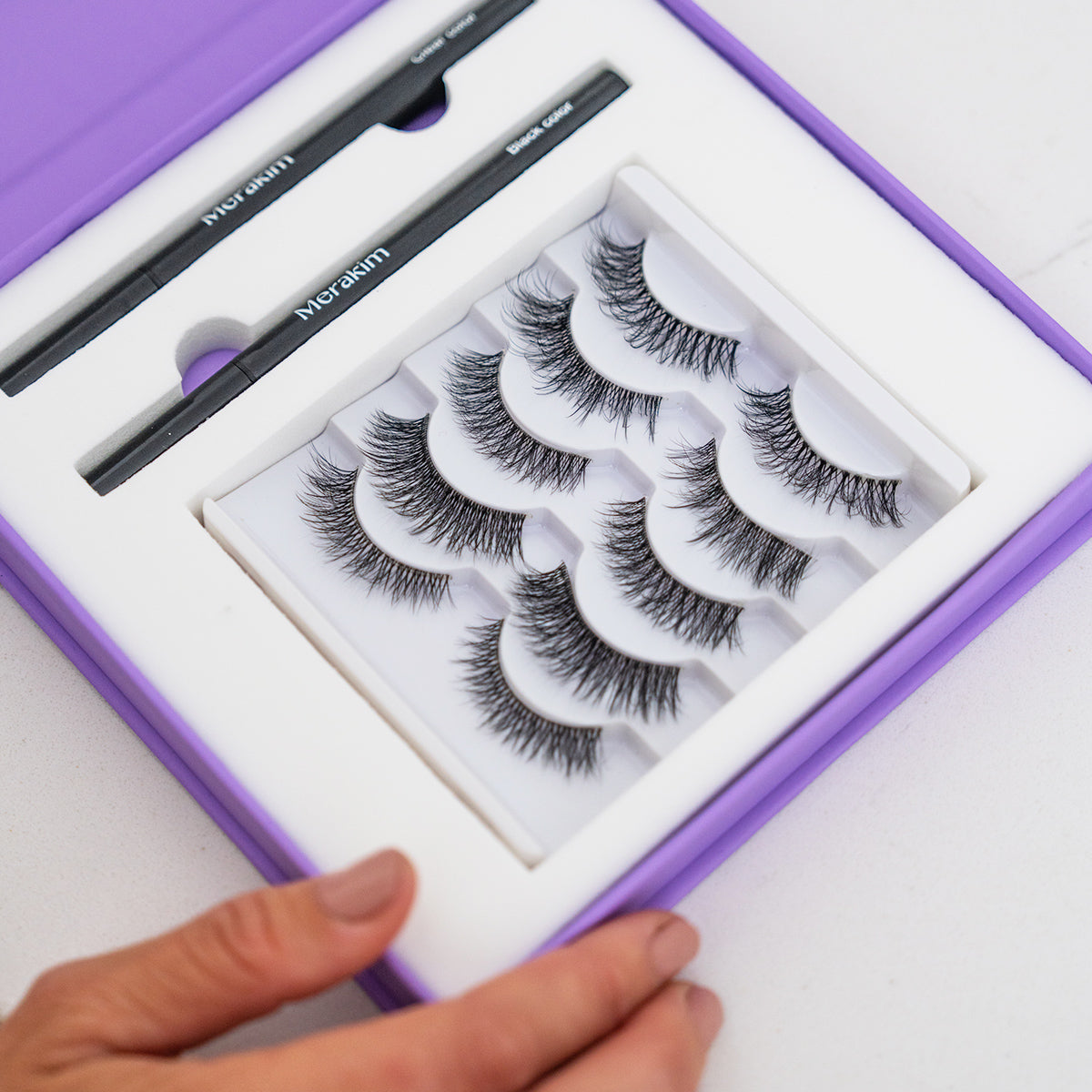 The Merakim Eyes on Beauty Lashes kit, an alternative to eyelash extensions, is elegantly displayed in a purple box with five pairs of lashes and two black magnetic eyeliners as a hand opens it on a white surface.