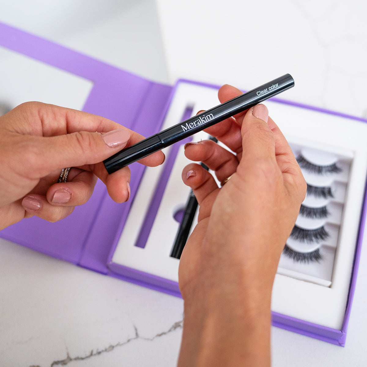 A person holds a Merakim magnetic eyeliner pen above an open purple box containing the Eyes on Beauty Lashes kit (5 pairs of faux mink lashes) on a pristine white surface.