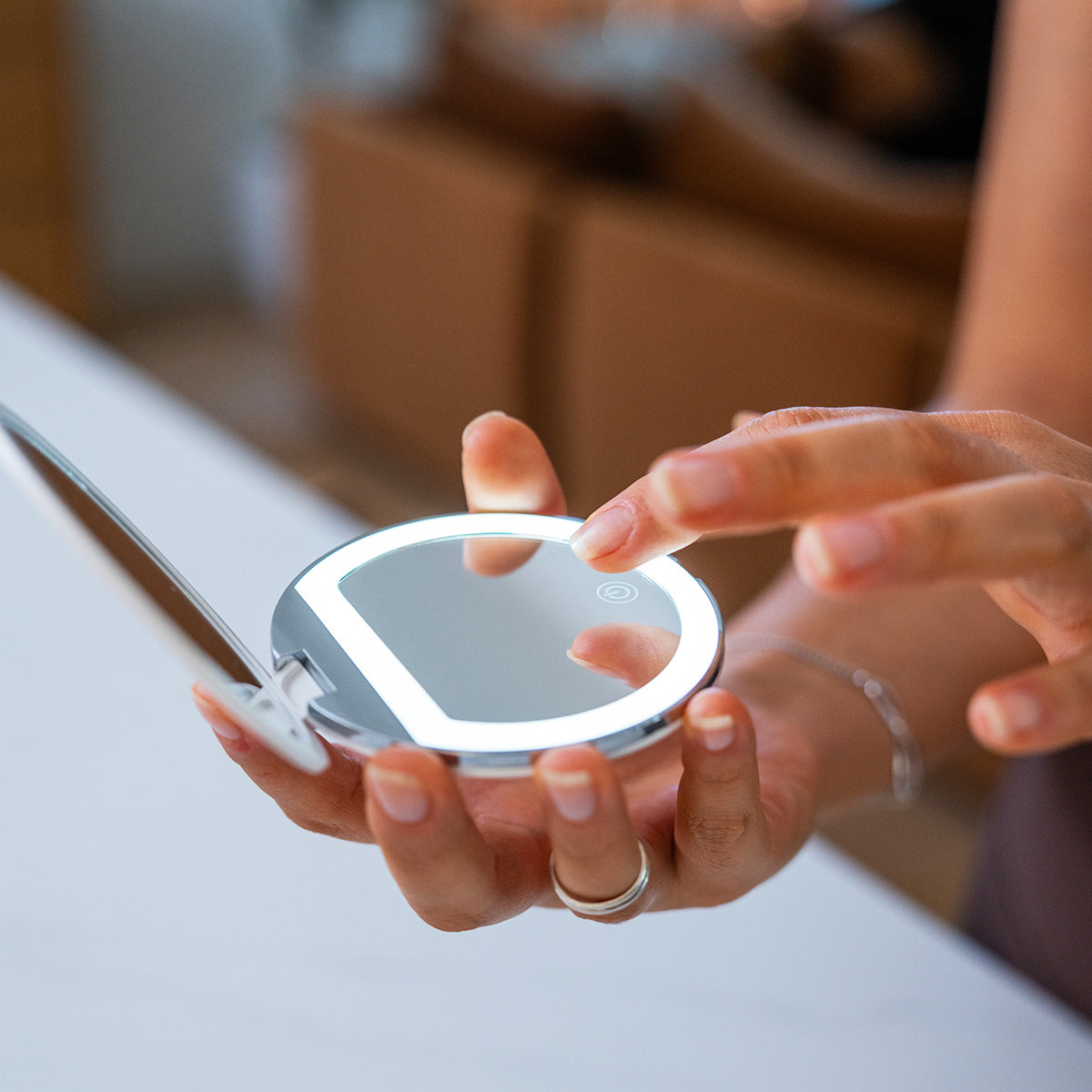 A close-up shot features a person holding the Merakim Rechargeable LED Makeup Mirror, showcasing its compact round design with illuminated LED ring. Their fingers touch the mirrors surface as they adjust the light settings, all set against a blurred background for focus.