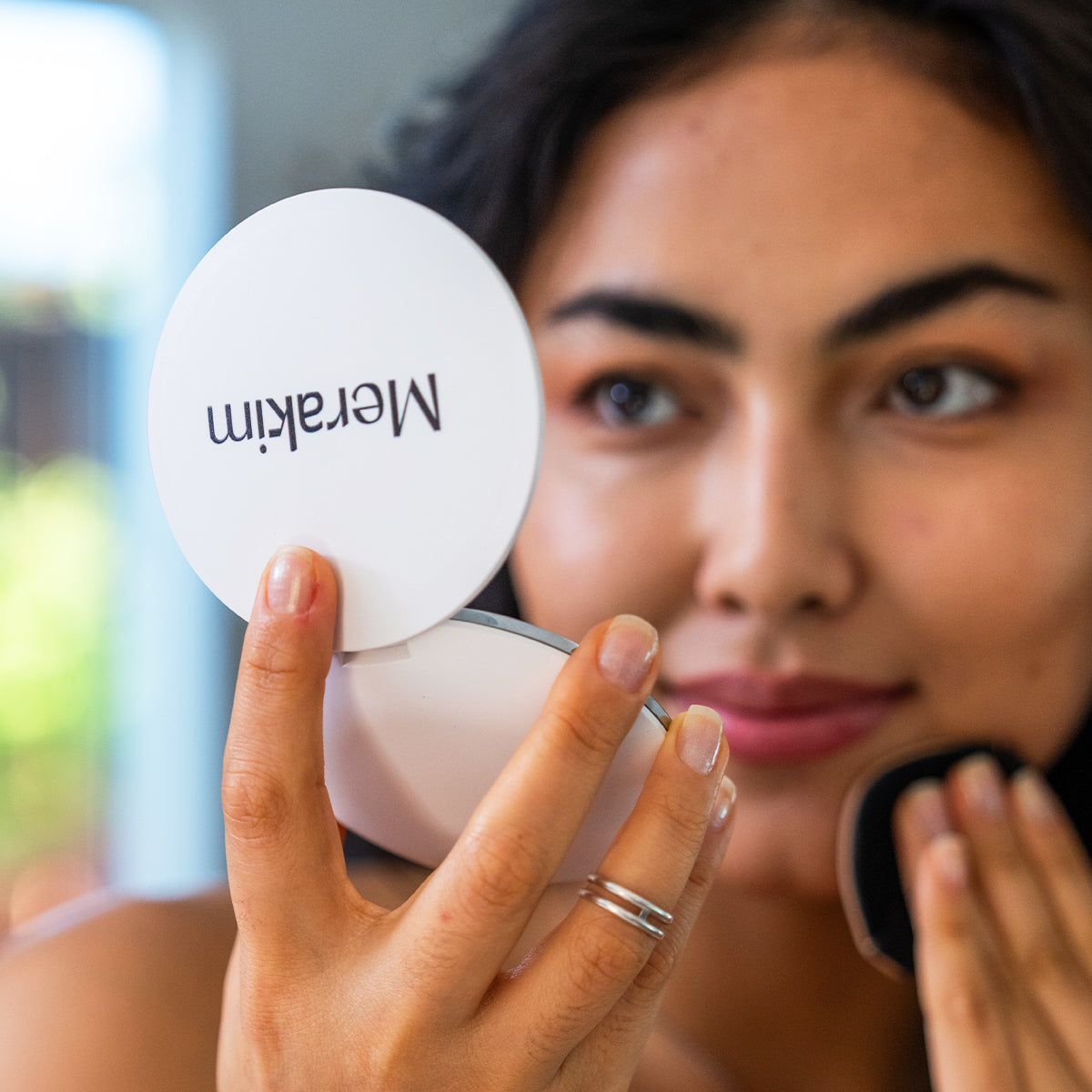 A person applies makeup using a Merakim Rechargeable LED Makeup Mirror, its adjustable light illuminating their face. The mirror features Merakim, and they wear a silver ring, smiling slightly. The softly blurred background suggests an indoor setting.