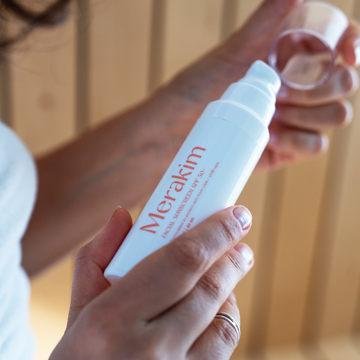 Close-up of a hand holding a white bottle labeled Merakim Facial Sunscreen SPF 50, ideal for sensitive skin, with the other hand removing the cap. Wooden vertical slats in the background highlight care and protection against UVA and UVB rays.