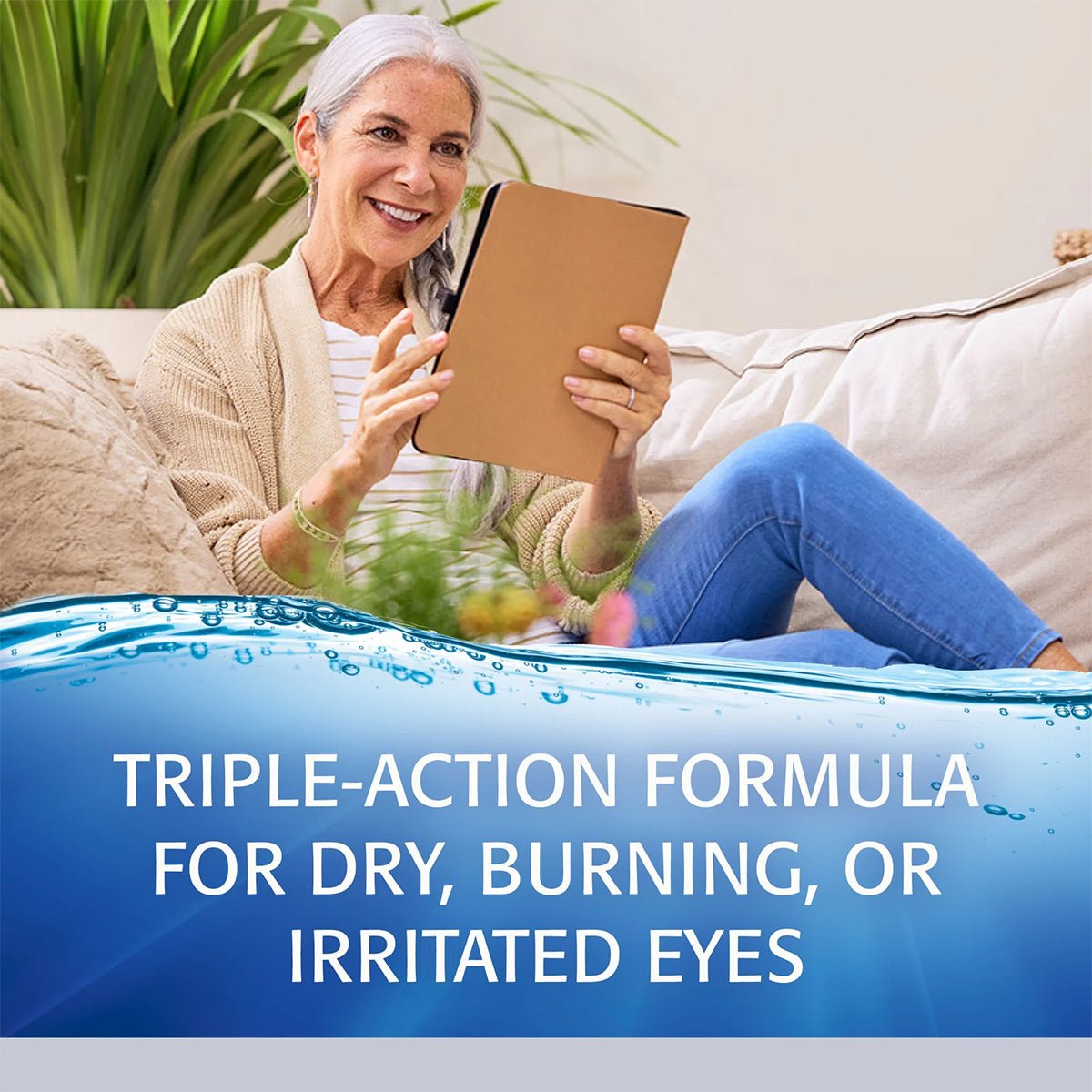 An older woman, wearing a beige cardigan and blue pants, sits on a couch smiling with Abbvies Refresh Optive Advanced Eye Drops (10 mL bottle), which offers HydroCell™ Technology for triple-action relief from dry, burning, or irritated eyes.