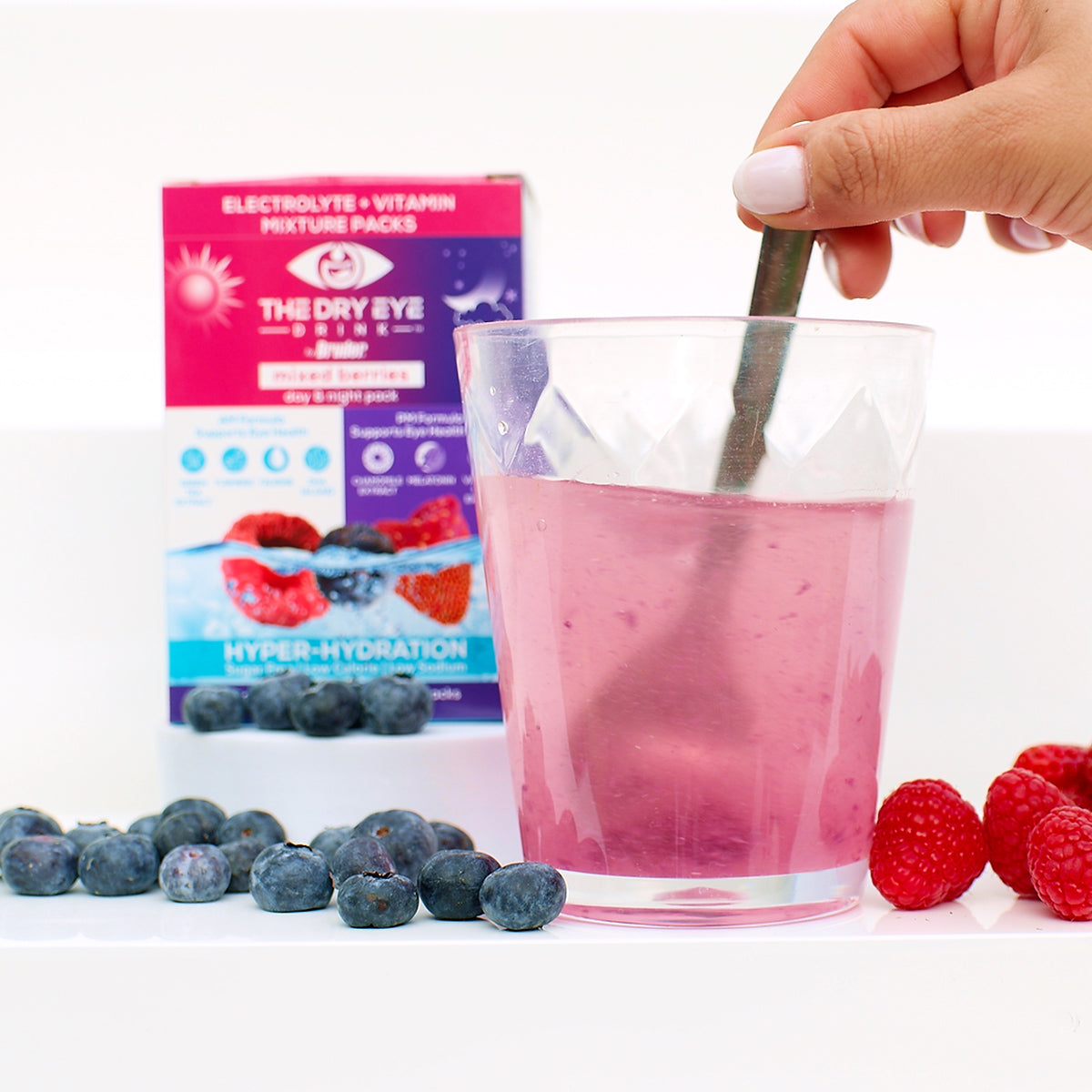 A hand stirs a glass of pink Dry Eye Drink from Bruder, surrounded by scattered blueberries and raspberries. In the background, a purple and pink box labeled Dry Eye Drink™ Complete Set offers hydration with electrolyte and vitamin packs for eye health.