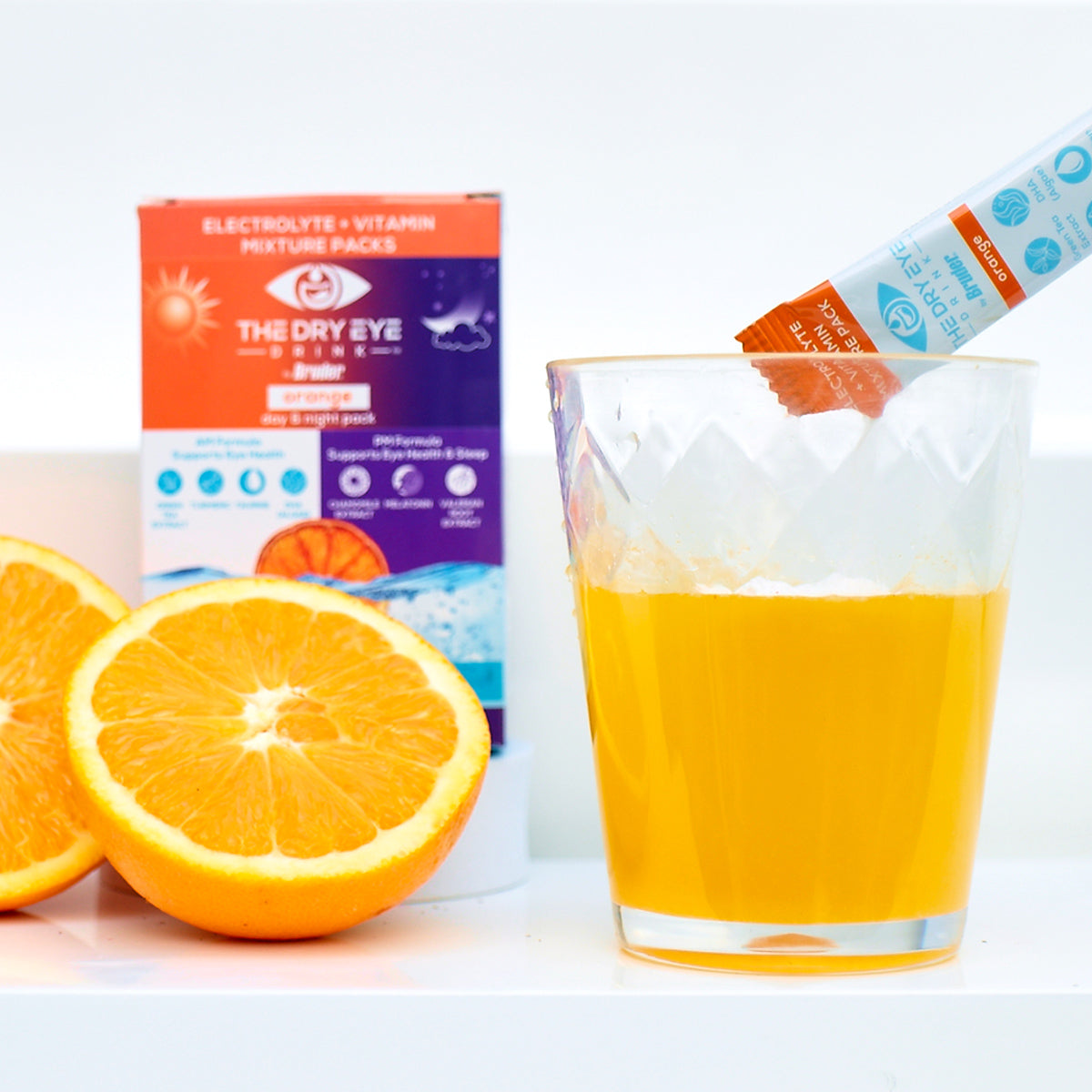 A glass with an orange drink sits beside a sliced orange, alongside the Dry Eye Drink™ Complete Set – Daytime & PM Hydration for Dry Eyes, promoting hydration for dry eyes. A branded Bruder box highlights electrolytes and vitamins for eye health.