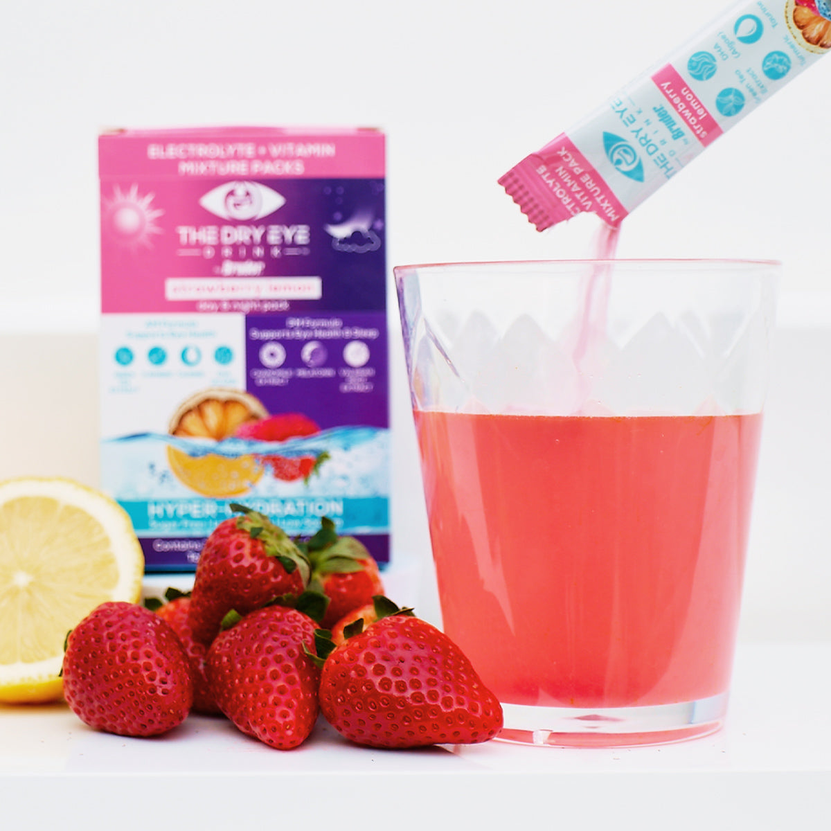 A pink electrolyte drink is elevated as a Dry Eye Drink™ packet from Bruder dissolves. Fresh strawberries and a lemon slice tempt in front, while the product packaging promises hydration and dry eye relief.