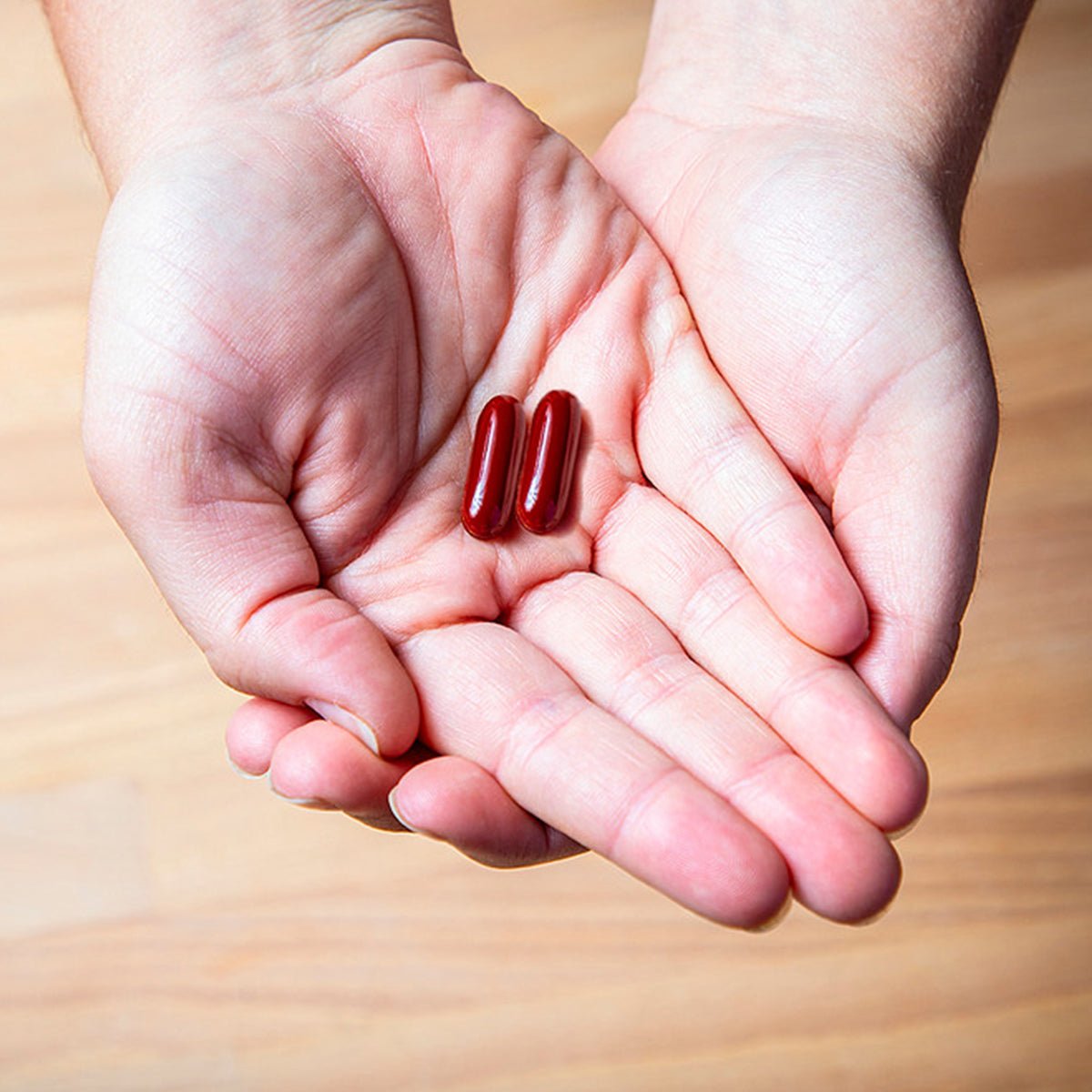 Two red capsules of EyePromise Macular Health Complete AREDS 2 based Multivitamin (60ct) sit in open palms against a wooden floor, representing vision support.