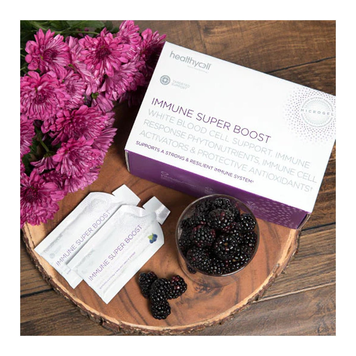 A box of HealthyCell Immune Support Boost Gel Packs with two sachets and a bowl of blackberries sits on a wooden table. Purple flowers and elderberries are arranged nearby, enhancing the natural aesthetic while supporting cell activation and your immune system.