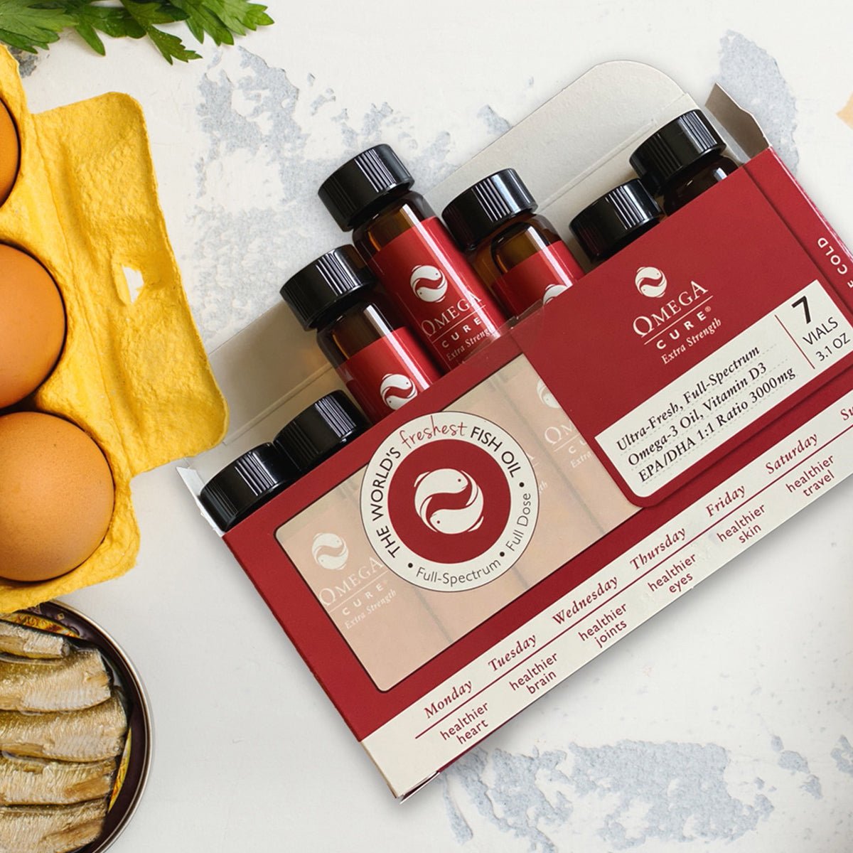 A flat lay features an Omega Cure Extra Strength Fresh Liquid Omega from Omega 3 Innovations, with seven vials partly inserted in a box. Nearby are brown eggs, a fish on a plate, and fresh green herbs. Cold shipping is included for the 1-week supply.