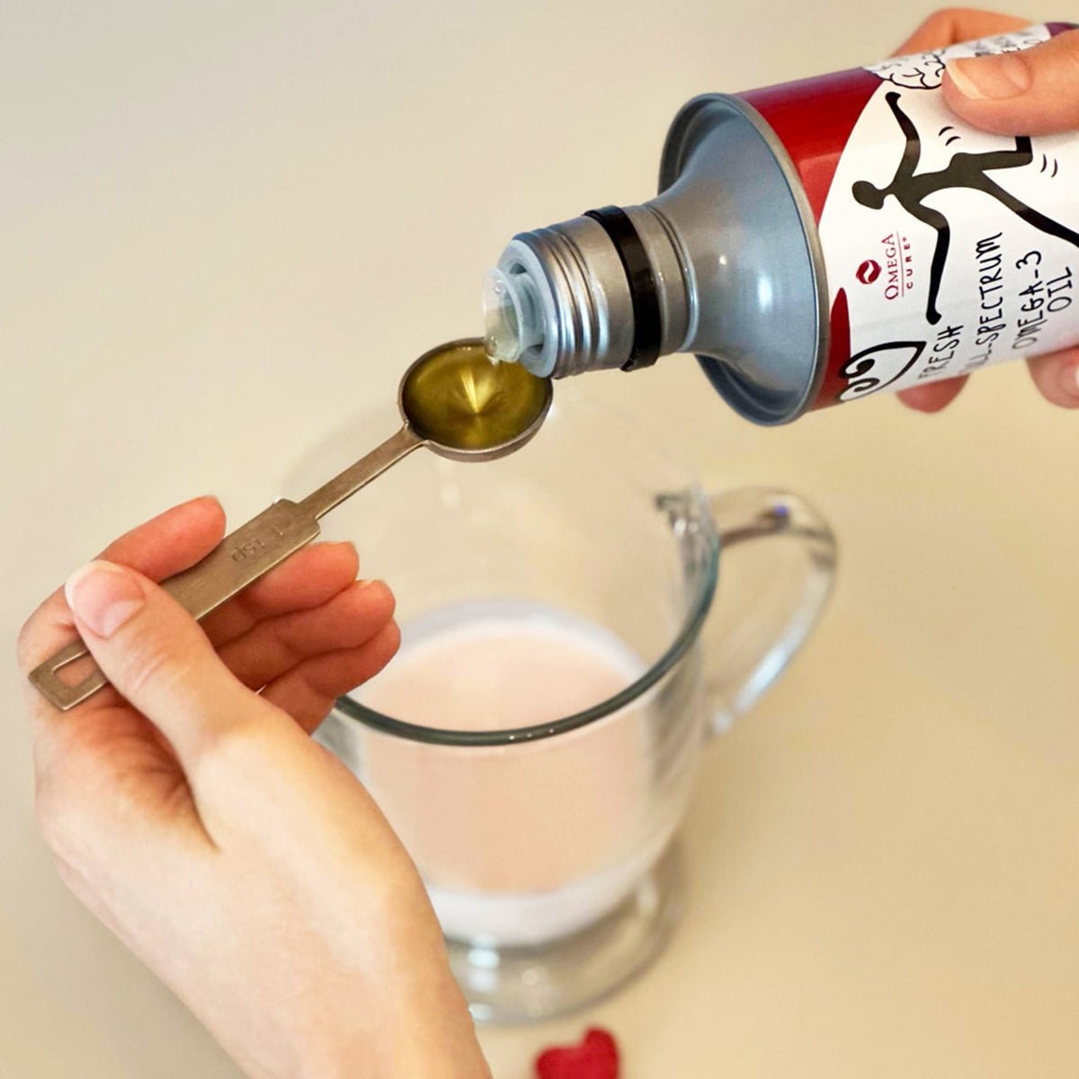 An individual pours Omega Cure Omega 3 Fish Oil from a bottle into a measuring spoon over a glass mug with pink liquid, on a beige surface. A small red heart cutout nearby symbolizes love for health and flexible dosing, highlighting the non-fishy taste of this Omega 3 Innovations product.