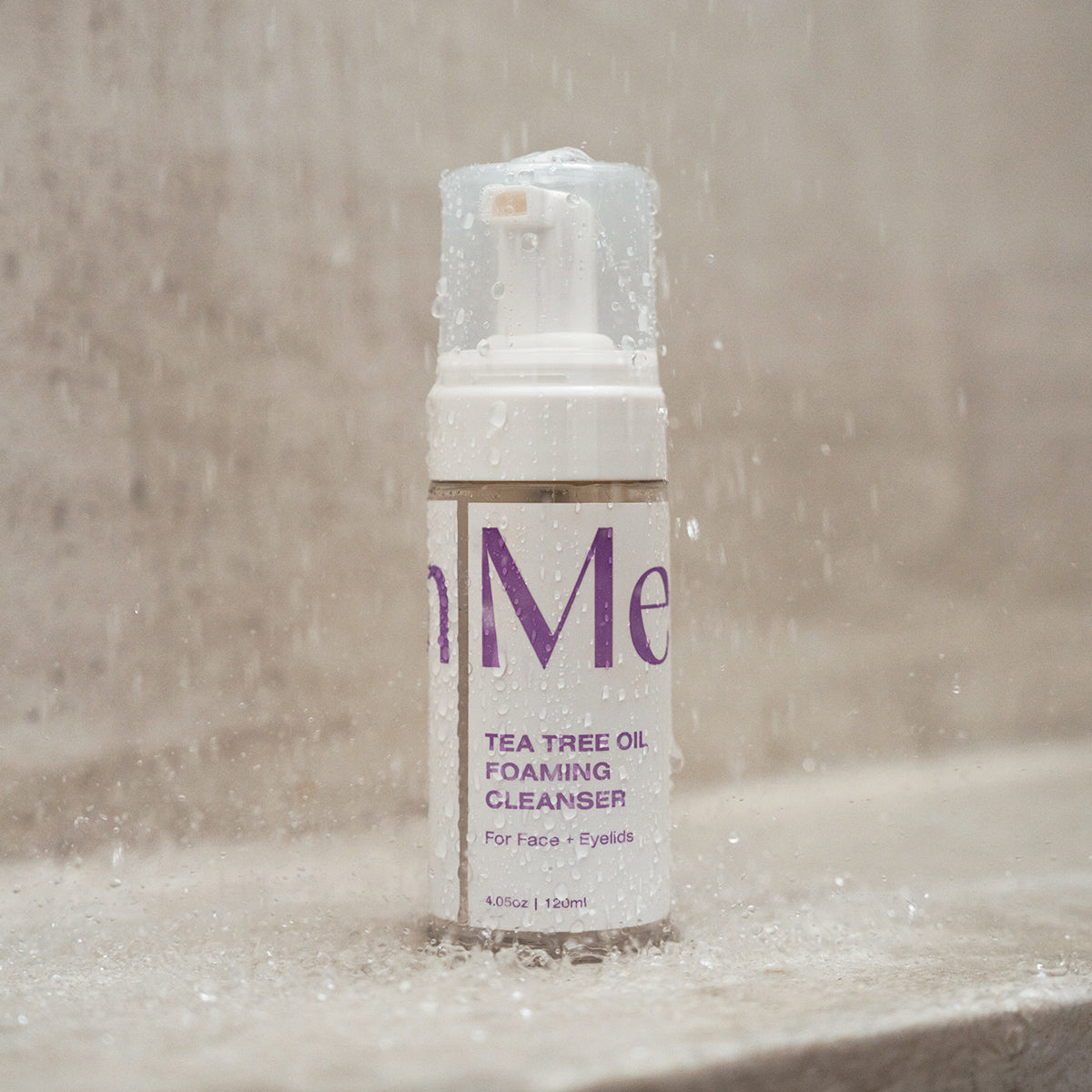 A Merakim Tea Tree Oil Foam Facial Cleanser (120mL) bottle stands upright with water droplets on a stone or tile surface, designed to soothe ocular inflammation. The label states its for face and eyelids, set against a softly blurred background.