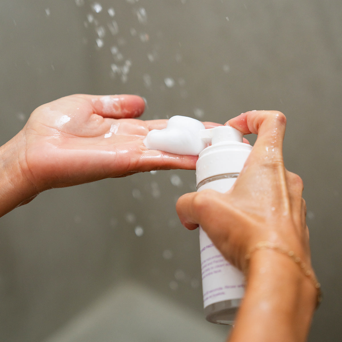 In the shower, a person dispenses Merakim Tea Tree Oil Foam Facial Cleanser (120mL) from a pump bottle onto their palm. Water droplets hover in the air, and their hands glisten slightly with moisture.