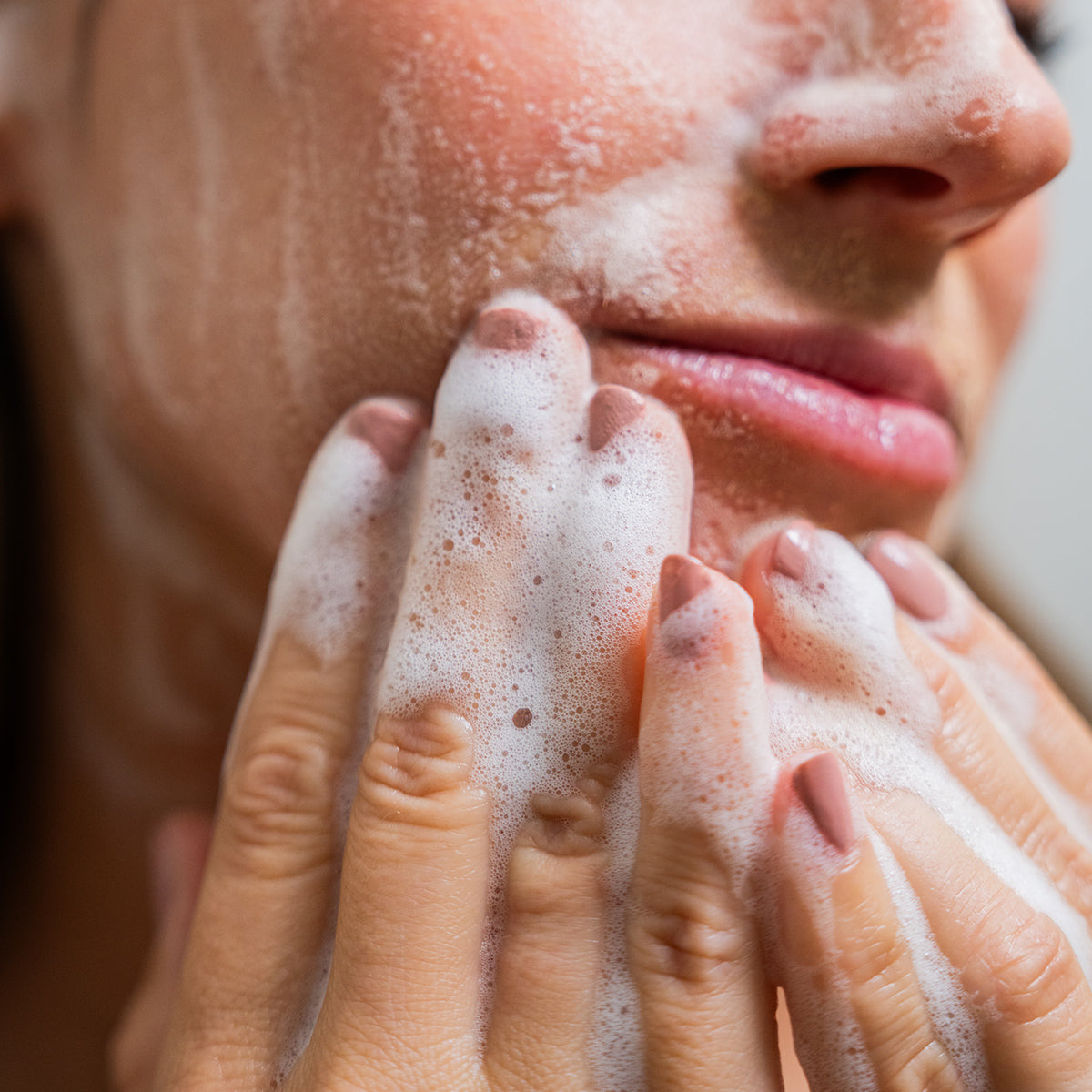 A close-up shows a person using Merakim Tea Tree Oil Foam Facial Cleanser (120mL), with hands covered in frothy soap gently massaging their cheeks. Their nails are painted in a neutral color, highlighting self-care and cleanliness.