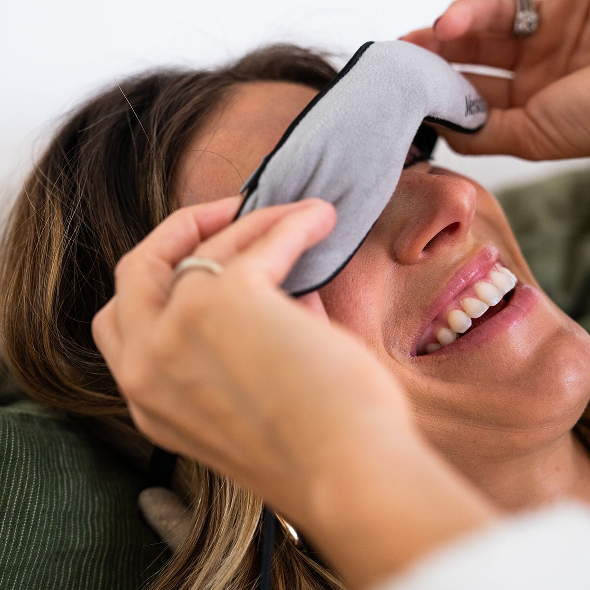 A woman smiles while adjusting a Merakim Professional USB Heat Eye Mask for dry eyes and tear film support, lying on a green cushion. Her hair is visible, and she wears a ring on her left hand, enjoying the soothing comfort of this adjustable warm compress.