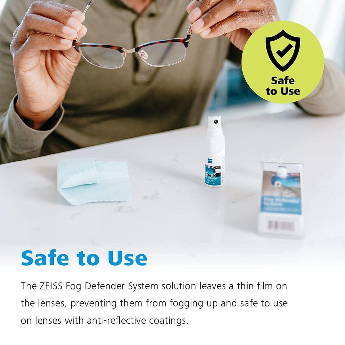 Someone applies ZEISS Fog Defender System Anti-Fog Spray by Zeiss to eyeglasses for fog-free vision. A microfiber cloth and packaging are on the table with a Safe to Use emblem, emphasizing the anti-fog solutions effectiveness for lenses with anti-reflective coatings.