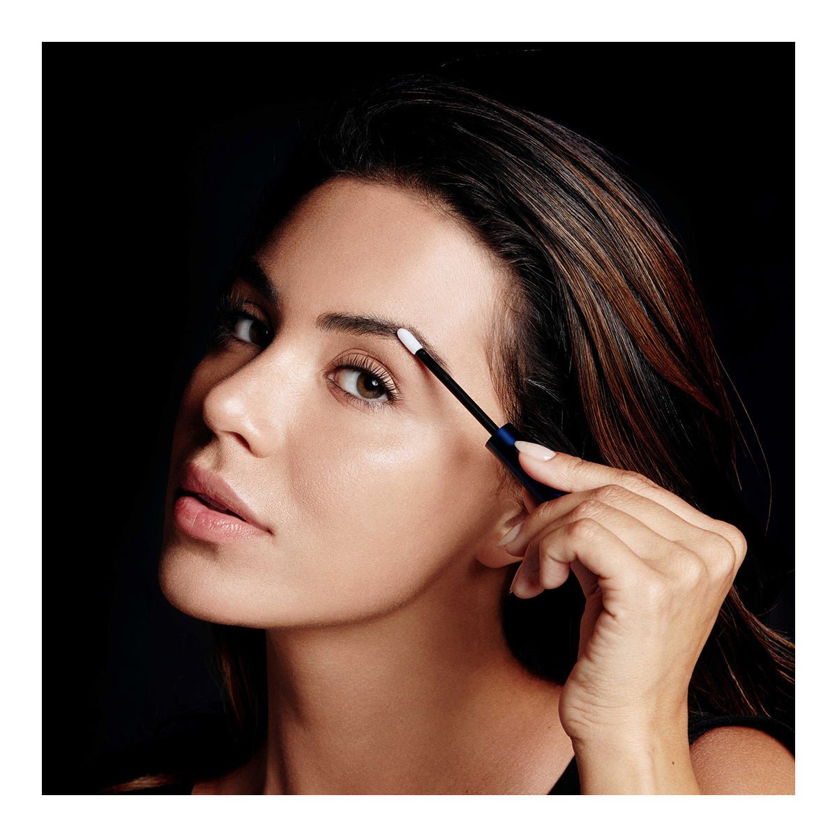 Against a black background, a person with dark hair neutrally applies RevitaLash RevitaBrow Advanced Eyebrow Conditioner Serum using a small brush, demonstrating the serums transformative effects and easy integration into any beauty routine.