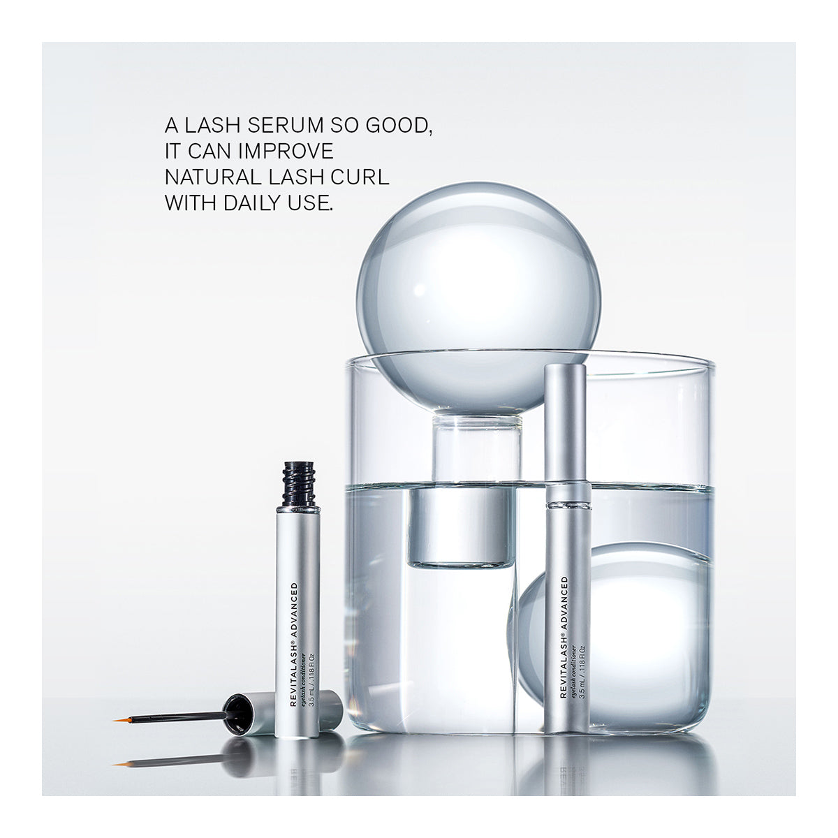 RevitaLash Advanced PRO Eyelash Conditioner and Enhancing Serum (3.5mL, 6 Mo. Supply) sits amid sleek metallic packaging and glass spheres filled with water. Text reads, A lash-enhancing formula so good, it can improve natural lash curl with daily use.