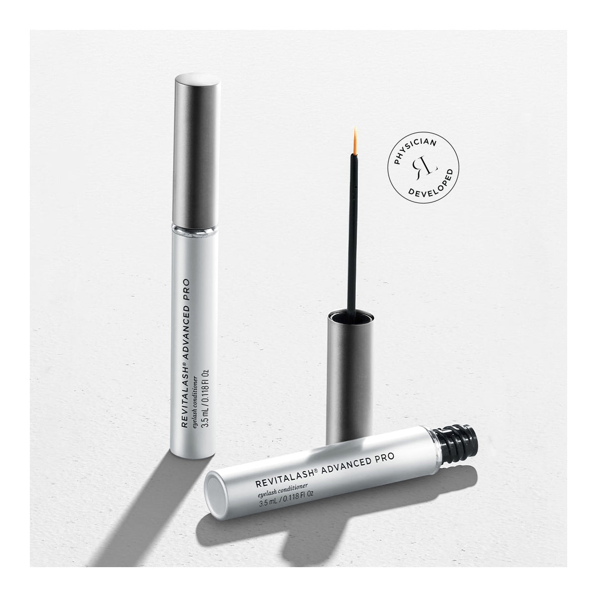 Two tubes of RevitaLash Advanced PRO Eyelash Conditioner and Enhancing Serum are displayed—one closed, the other open with an applicator. The sleek white and silver packaging features Physician Developed elegantly inscribed, emphasizing its lash-enhancing properties.