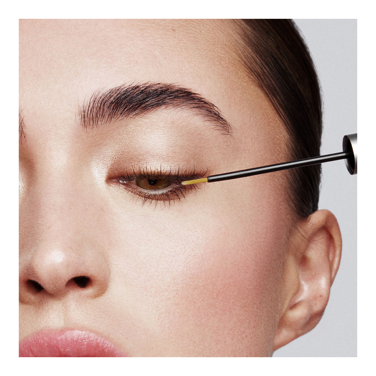 A close-up shows someone applying gold eyeliner on their upper eyelid, with well-groomed eyebrows and lashes enhanced by RevitaLash Advanced PRO Eyelash Conditioner and Enhancing Serum. The eye is partially closed against a plain white background, highlighting the subtle makeup.