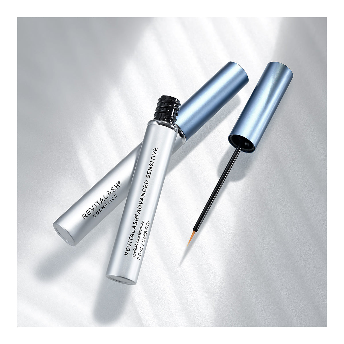 A blue tube of RevitaLash Advanced Eyelash Conditioner and Enhancing Serum Sensitive (2.0mL 3 Mo. Supply) with its cap off shows an applicator brush resting on the opening. The product, promoting lash health, stands against a soft, white textured background.