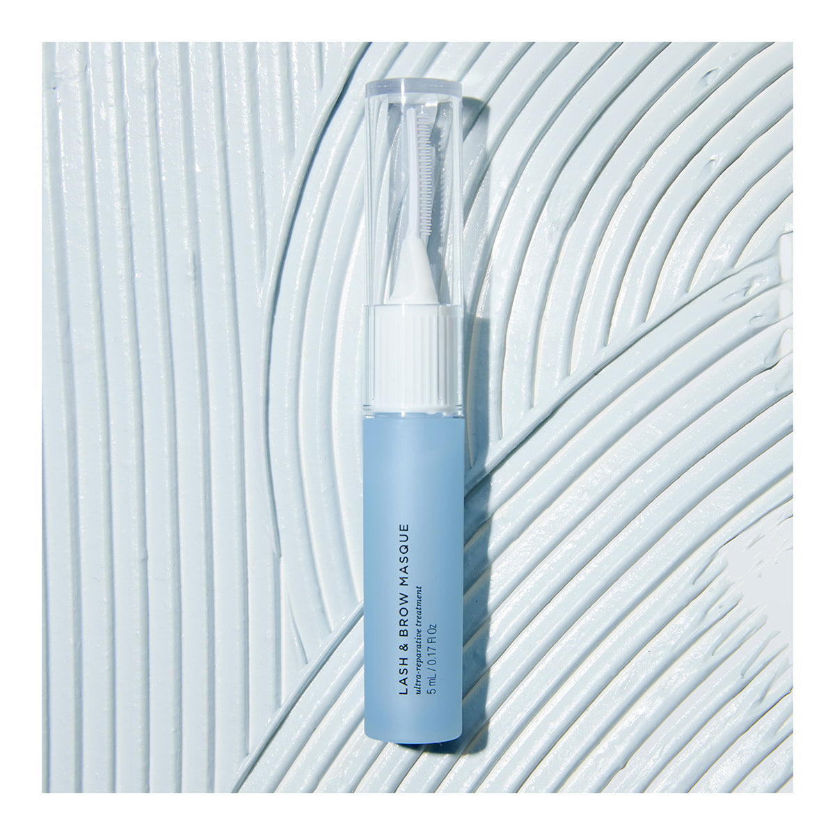 A blue tube of RevitaLash Lash and Brow Masque with a translucent cap lies on a textured, light blue surface. The tube displays white text highlighting the product name and RevitaLash branding.