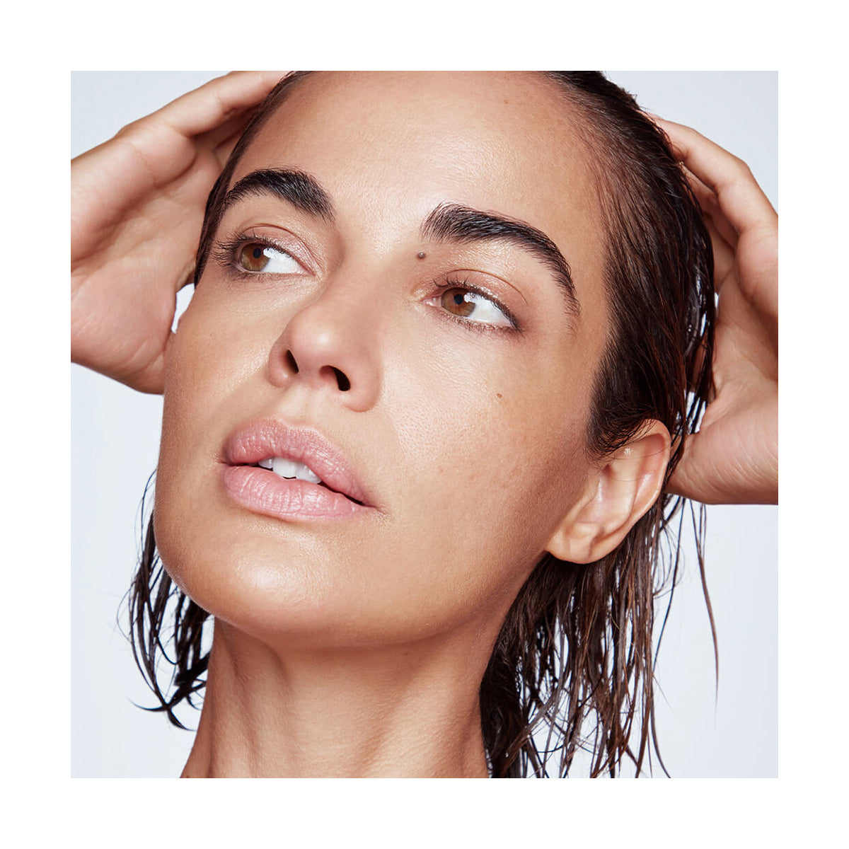 With wet hair treated by RevitaLashs Thickening Conditioner Scalp Therapy Formula (250mL), hypoallergenic and neutral against a light plain background, they tilt their head slightly, holding it with both hands and gaze distantly.