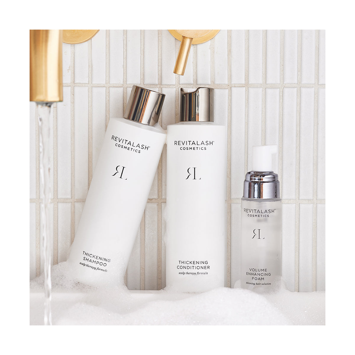 Three RevitaLash Volumizing Hair Collection Bundle items—thickening shampoo, conditioner, and foam—sit on a tiled bathroom surface with running water and bubbles.