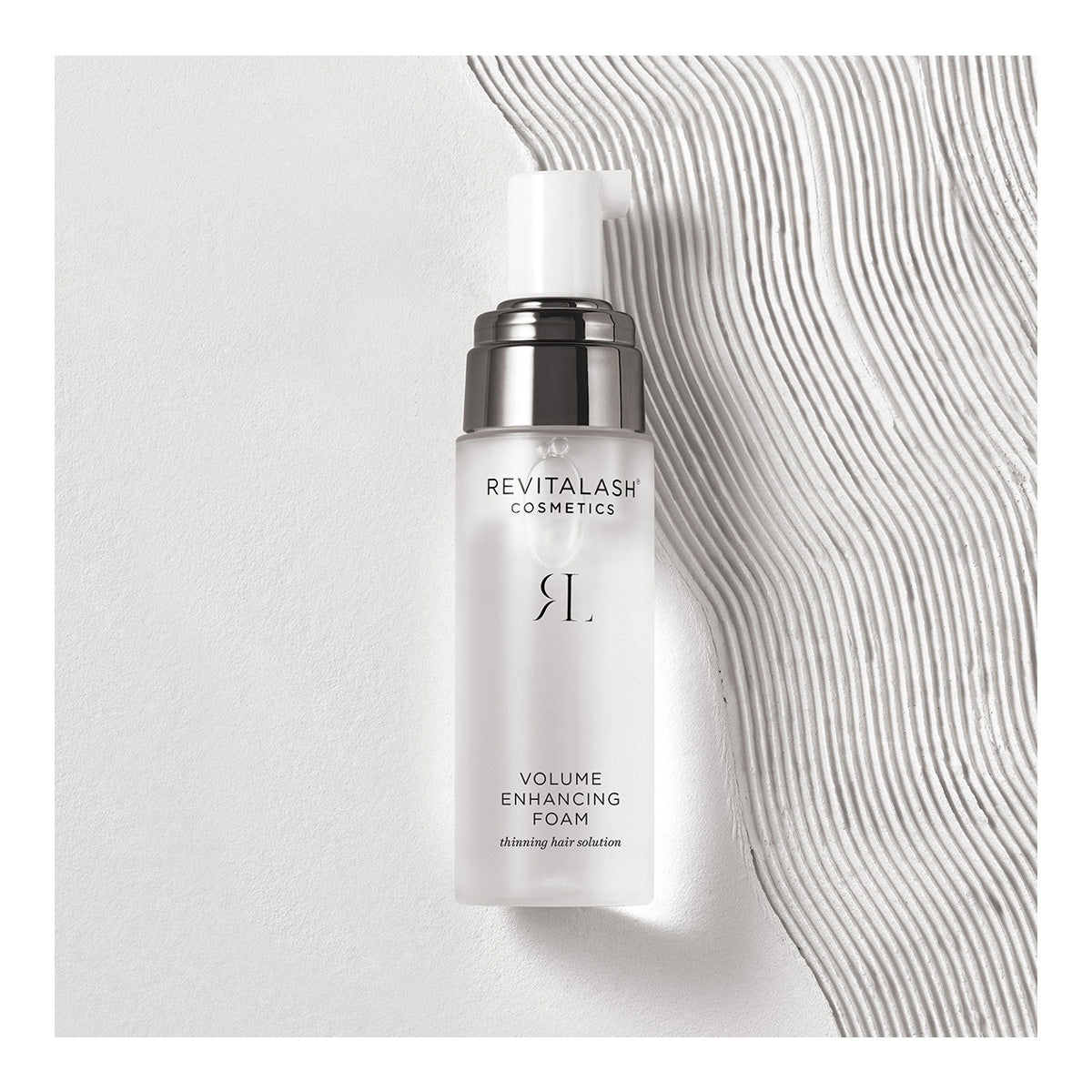 A bottle of RevitaLash Volume Enhancing Foam for Hair (55mL) with its elegant frosted pump dispenser rests on a textured white surface. Its minimalist design, subtle branding, and clear text emphasize its role as a luxurious hair enhancer.