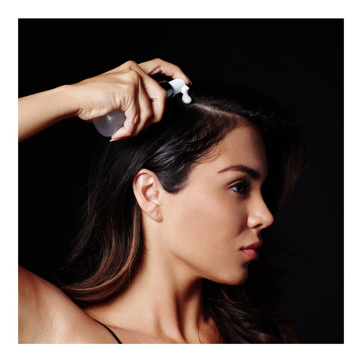 A person applies RevitaLash Volume Enhancing Foam for Hair (55mL) to dark hair, focusing on the hairline. The black background highlights skin and defined features, while the concentrated expression reflects dedication to its BioPeptin Complex benefits.