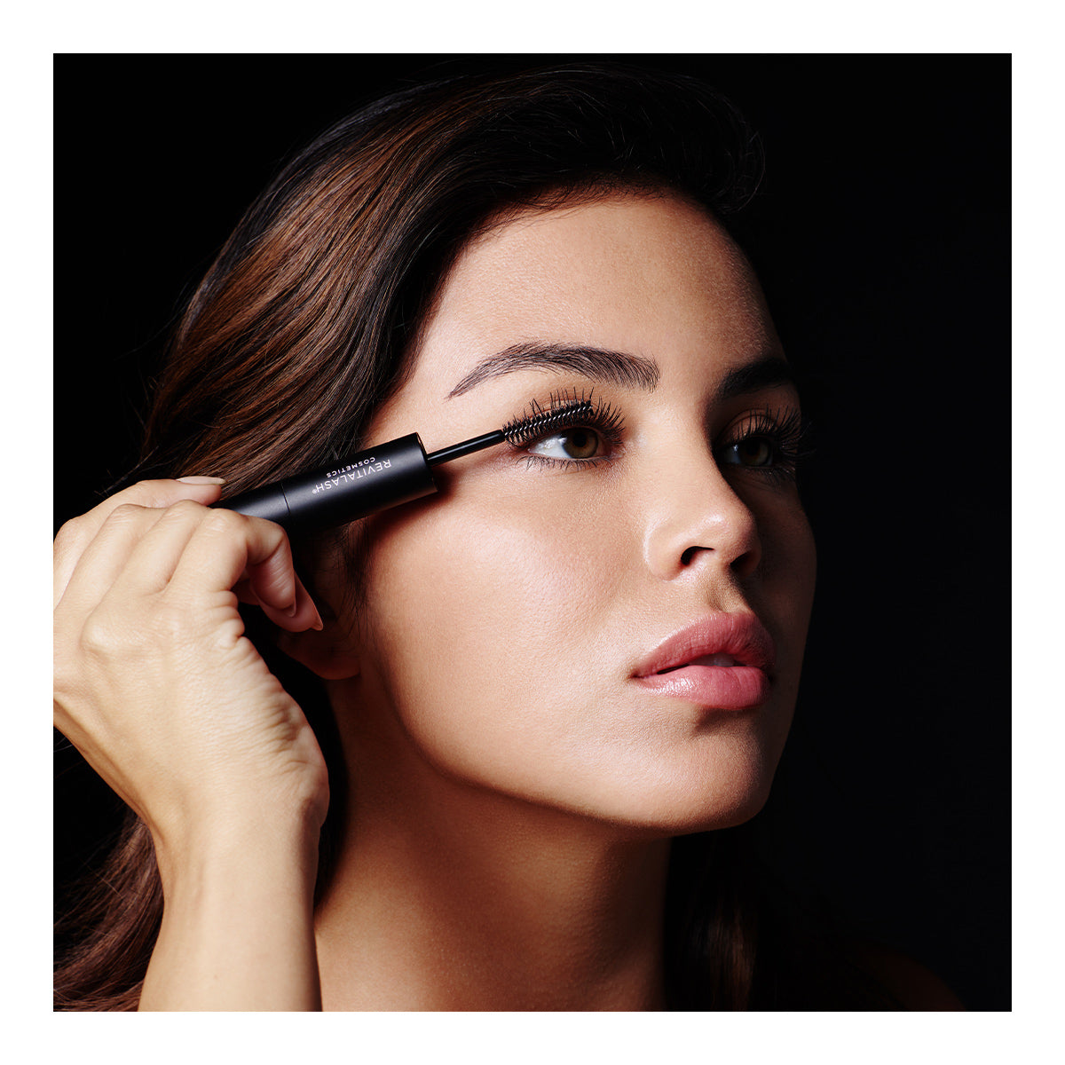 A woman with long, dark hair applies RevitaLash Double-Ended 2-in-1 Lash Primer & Mascara Volume Set to her eyelashes. She looks upward against a dark background. Her subtle makeup highlights her eyes and lips. With a serious expression, she embodies the elegance of RevitaLash.