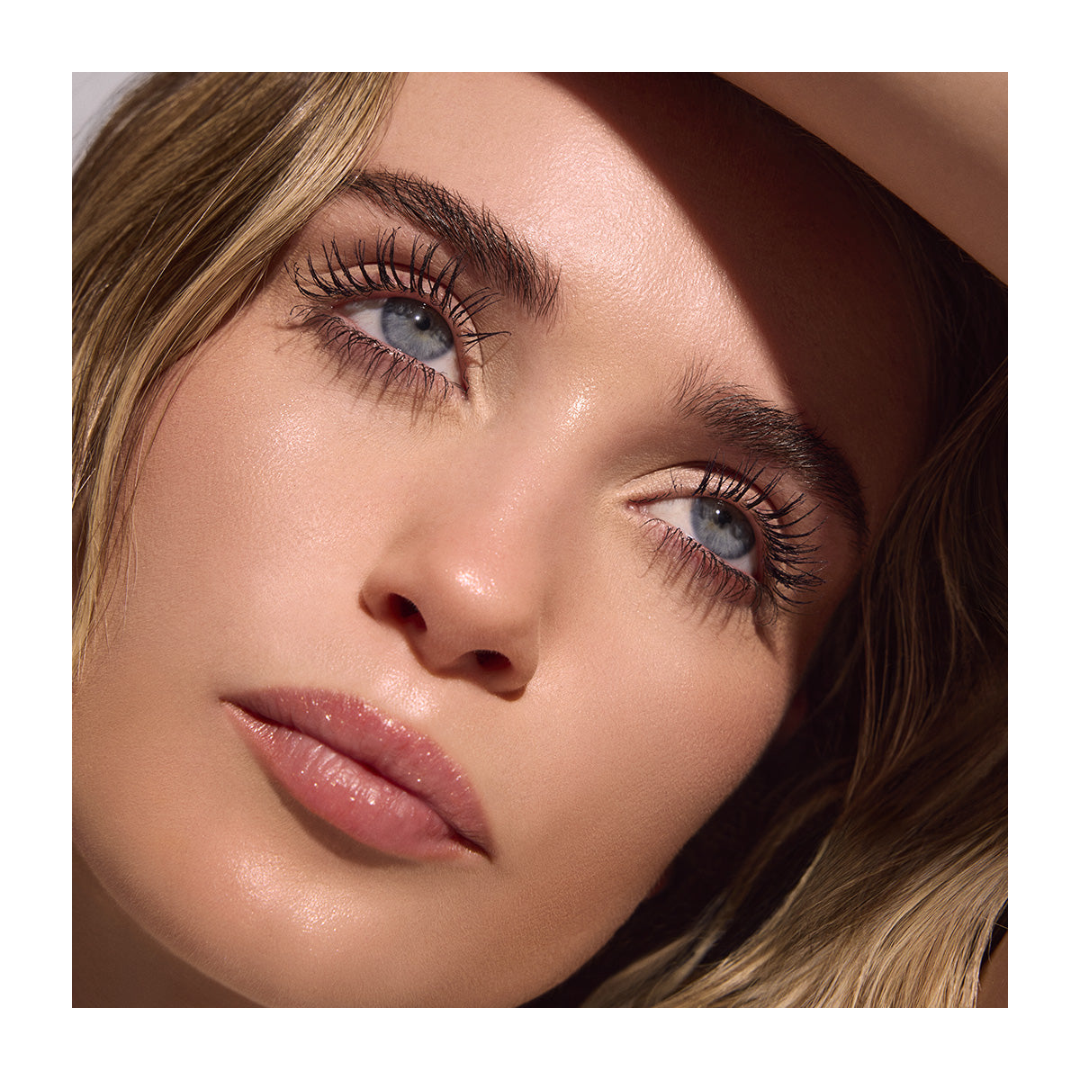 A close-up shows a person with light skin and blue eyes, wearing RevitaLash Length Define Tubing Mascara on long, dark lashes. With subtle makeup and light blonde hair, they gaze upward softly as delicate shadows play on their face.