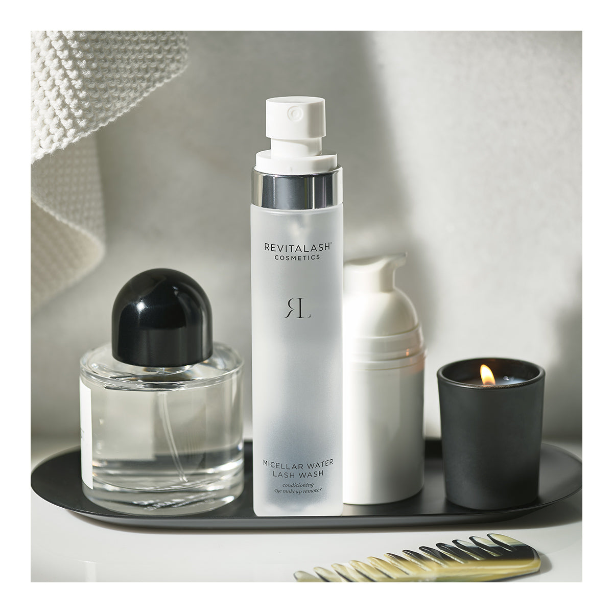 On a black tray, youll find the RevitaLash Micellar Water Lash Wash, a hypoallergenic makeup remover for sensitive eyes, alongside a Conditioning Eye Makeup Remover and a black and clear perfume bottle. Nearby sit a white foaming dispenser next to a lit candle, with a towel and hair comb adjacent.