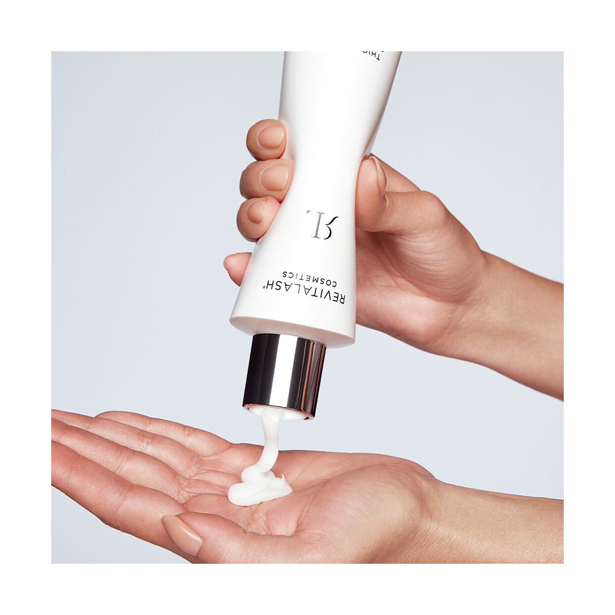 Hands squeezing RevitaLash Thickening Shampoo from a white tube with a silver cap and RL logo onto an open palm, ideal for scalp therapy. The background is light-colored and plain.