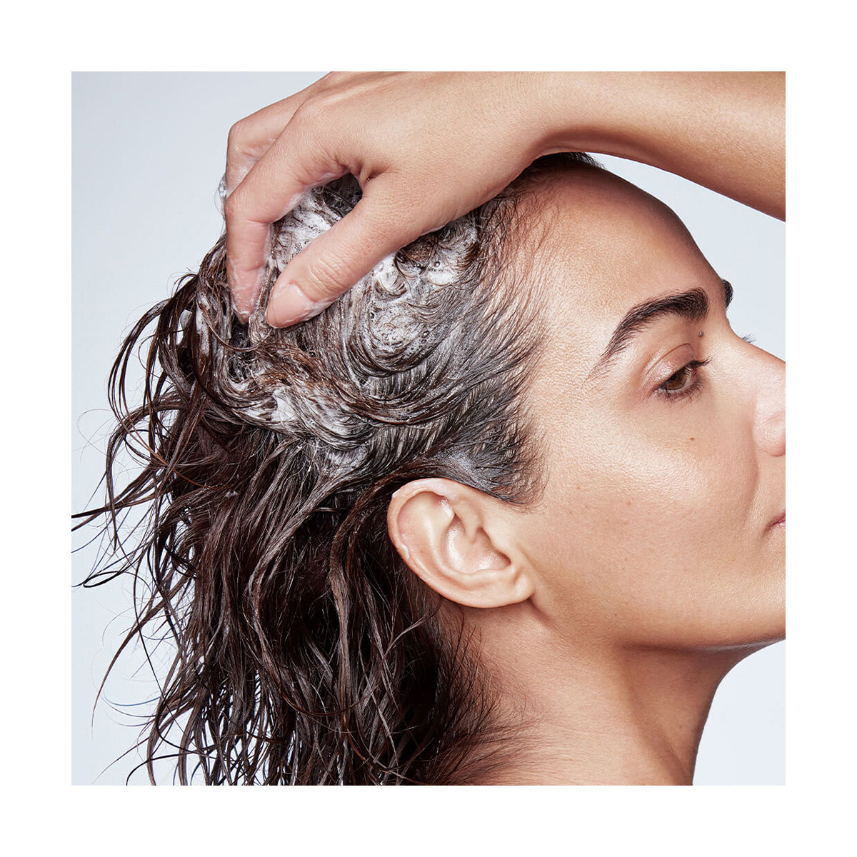 A person uses RevitaLash Thickening Shampoo, lathering it into their scalp with one hand. The wet hair partially covers their ear, highlighting a focus on hair health, as seen in a side profile against a light background.